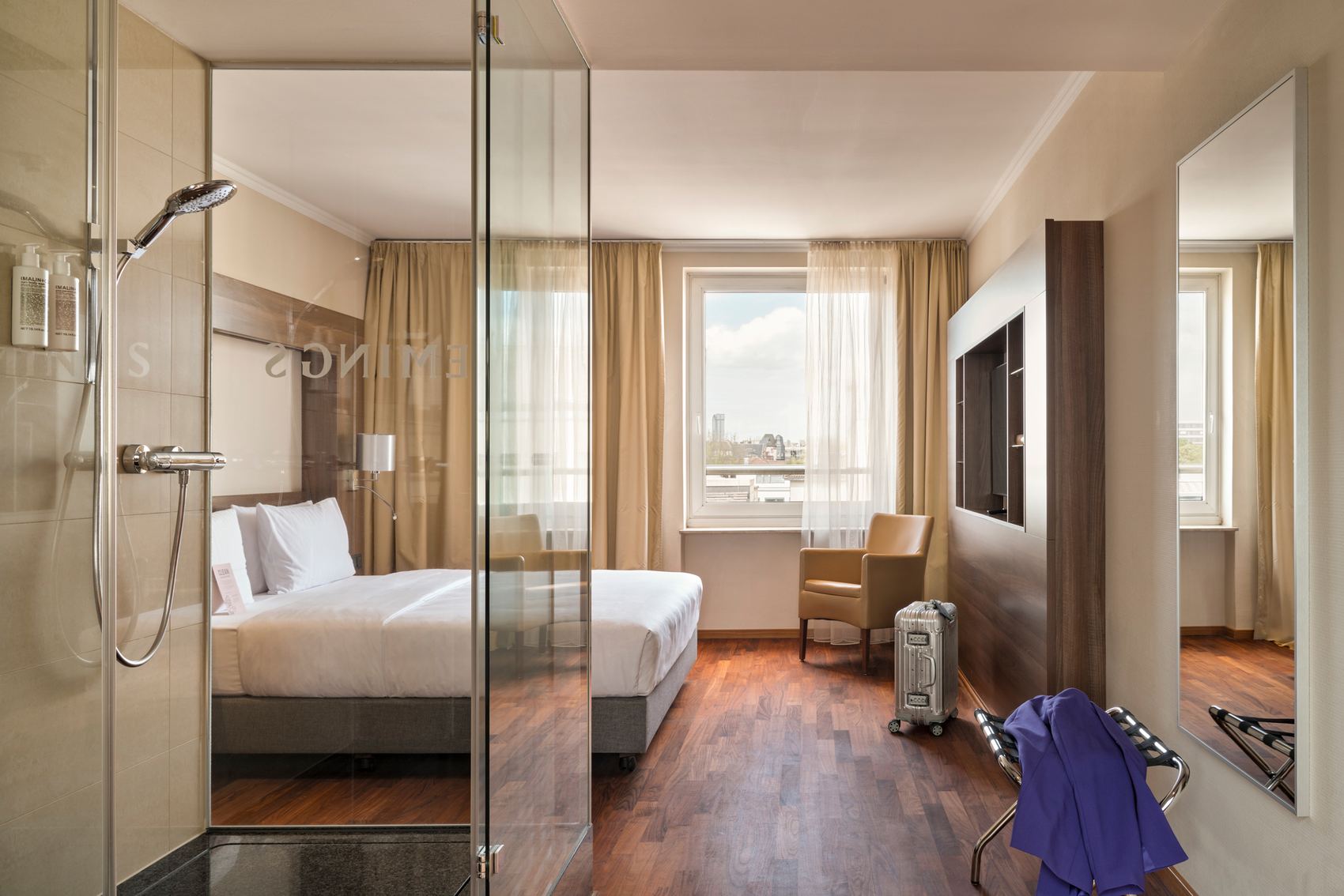Modernes Hotelzimmer mit großem Bett, Glasdusche, Holzfußboden und Tageslicht. Helle Vorhänge, bequemer Sessel am Fenster mit Stadtblick, minimalistische Einrichtung für entspannten Aufenthalt.