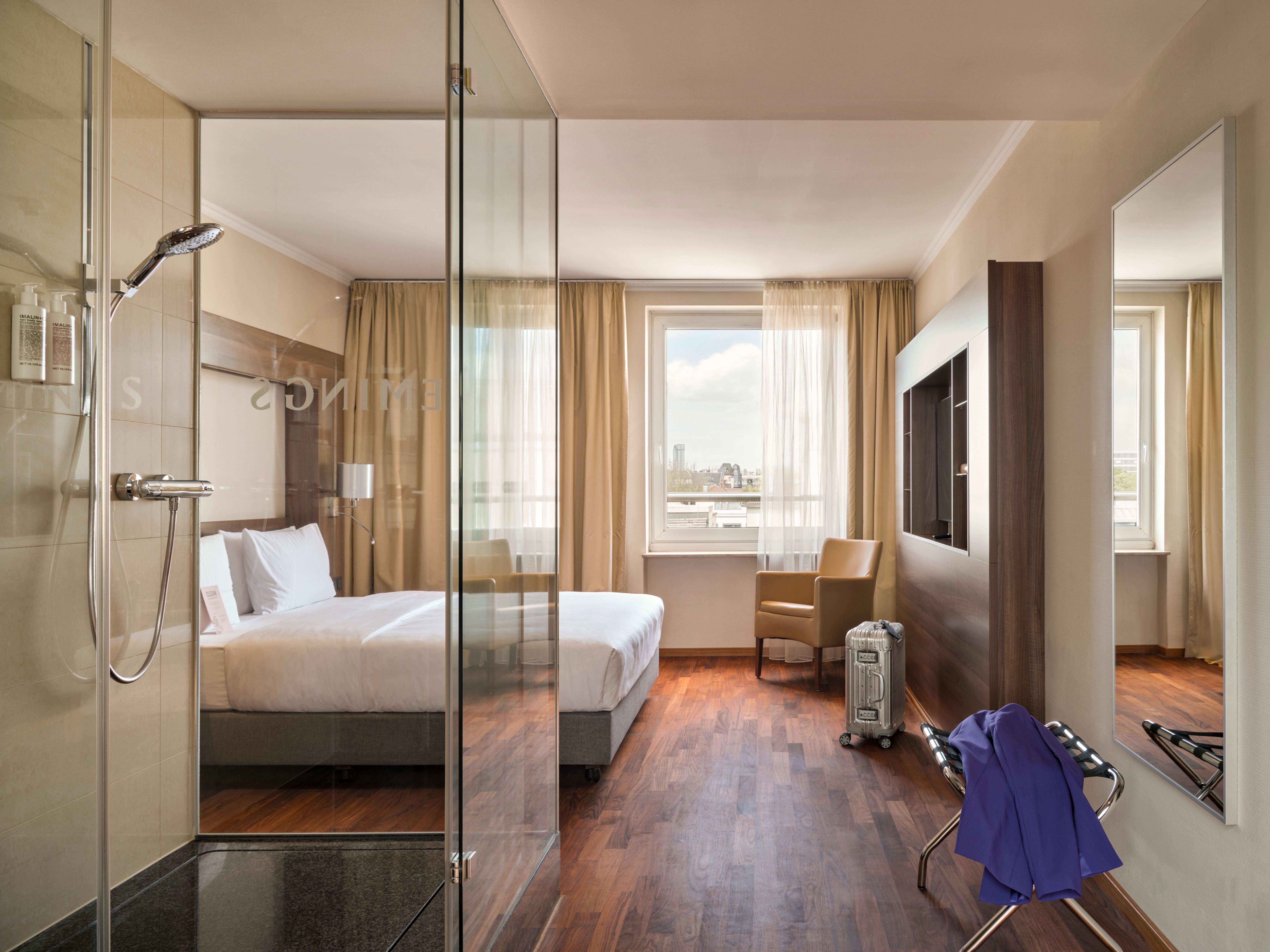 Modernes Hotelzimmer mit großem Bett, Glasdusche, Holzfußboden und Tageslicht. Helle Vorhänge, bequemer Sessel am Fenster mit Stadtblick, minimalistische Einrichtung für entspannten Aufenthalt. Modernes Hotelzimmer mit großem Bett, Glasdusche, Holzfußboden und Tageslicht. Helle Vorhänge, bequemer Sessel am Fenster mit Stadtblick, minimalistische Einrichtung für entspannten Aufenthalt.