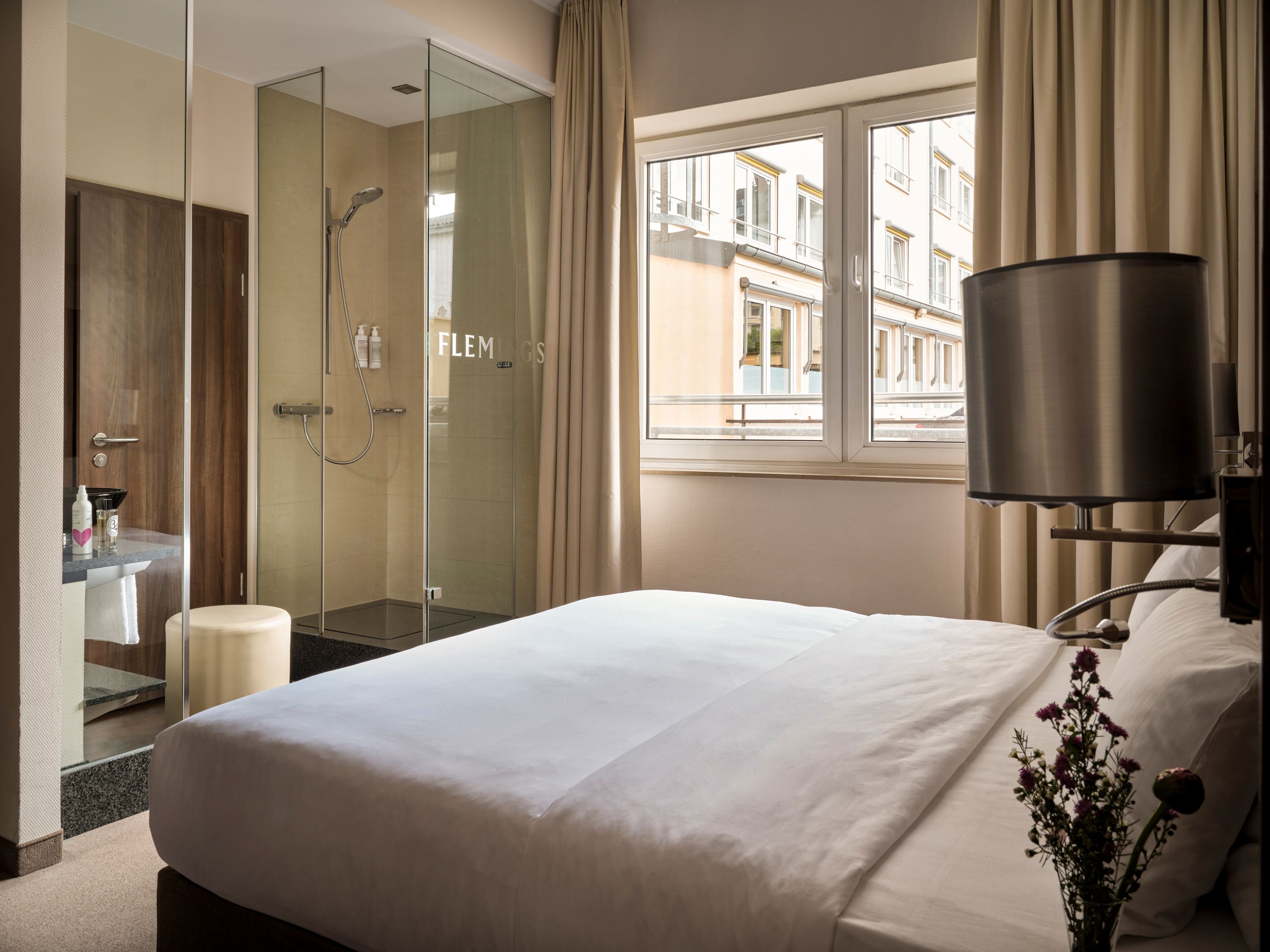Helles Hotelzimmer mit großem Bett, modernem Duschbereich aus Glas und Fenster mit Stadtblick, stilvoll eingerichtet für Komfort und Ruhe.