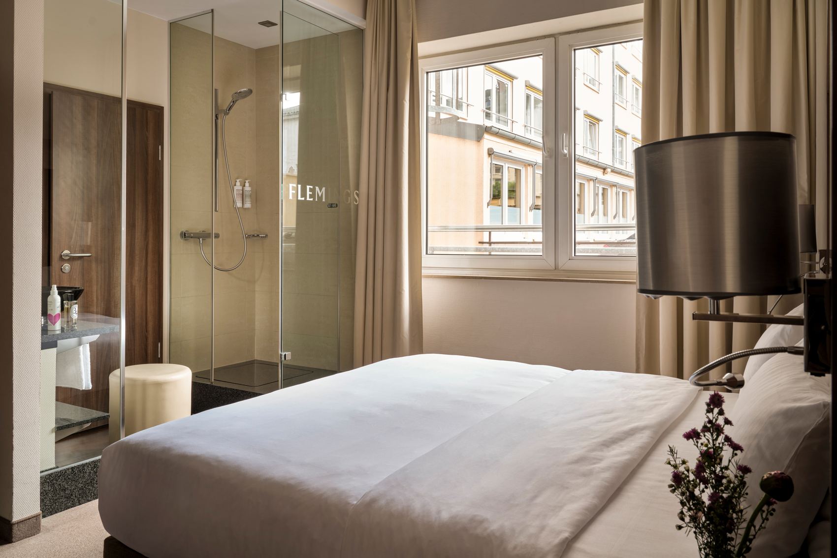 Helles Hotelzimmer mit großem Bett, modernem Duschbereich aus Glas und Fenster mit Stadtblick, stilvoll eingerichtet für Komfort und Ruhe.