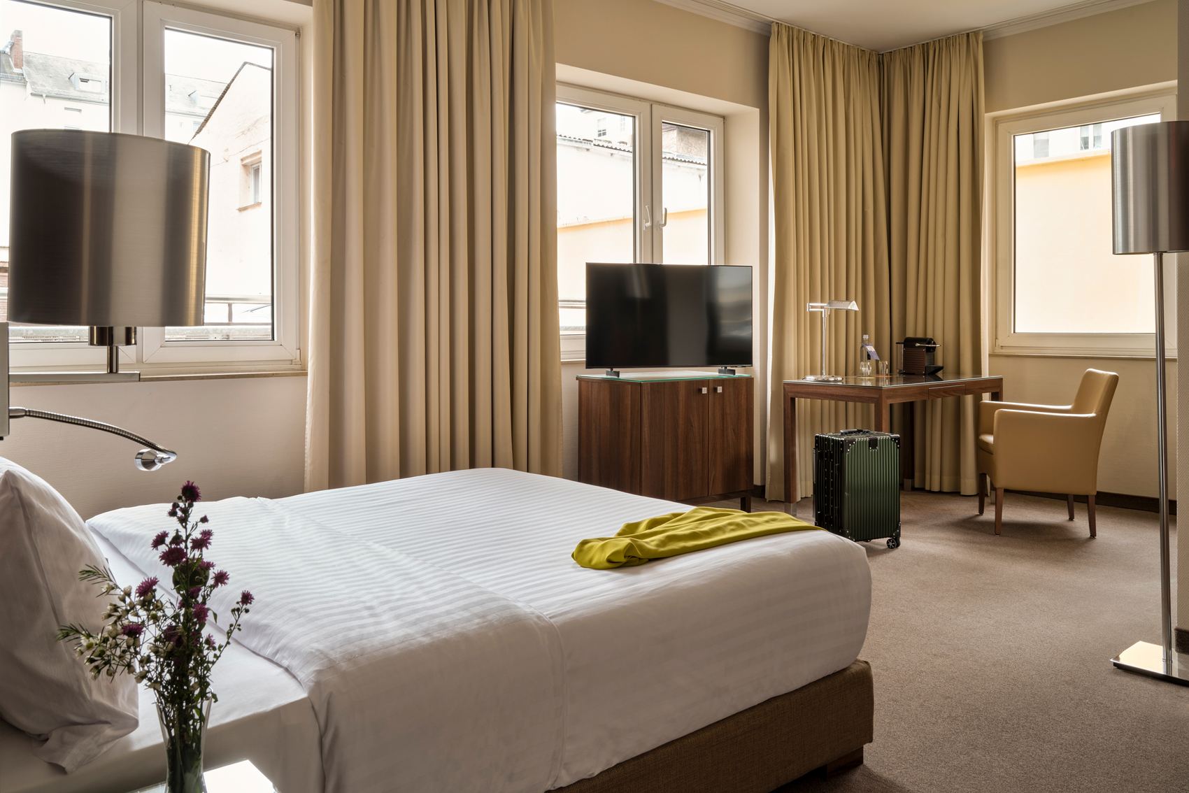 Helles Hotelzimmer mit großem Doppelbett, Fernseher, Schreibtisch und Sitzbereich. Drei Fenster bieten Tageslicht und Blick auf benachbarte Gebäude. Schlichte, warme Farben und moderne Einrichtung schaffen eine angenehme Atmosphäre.