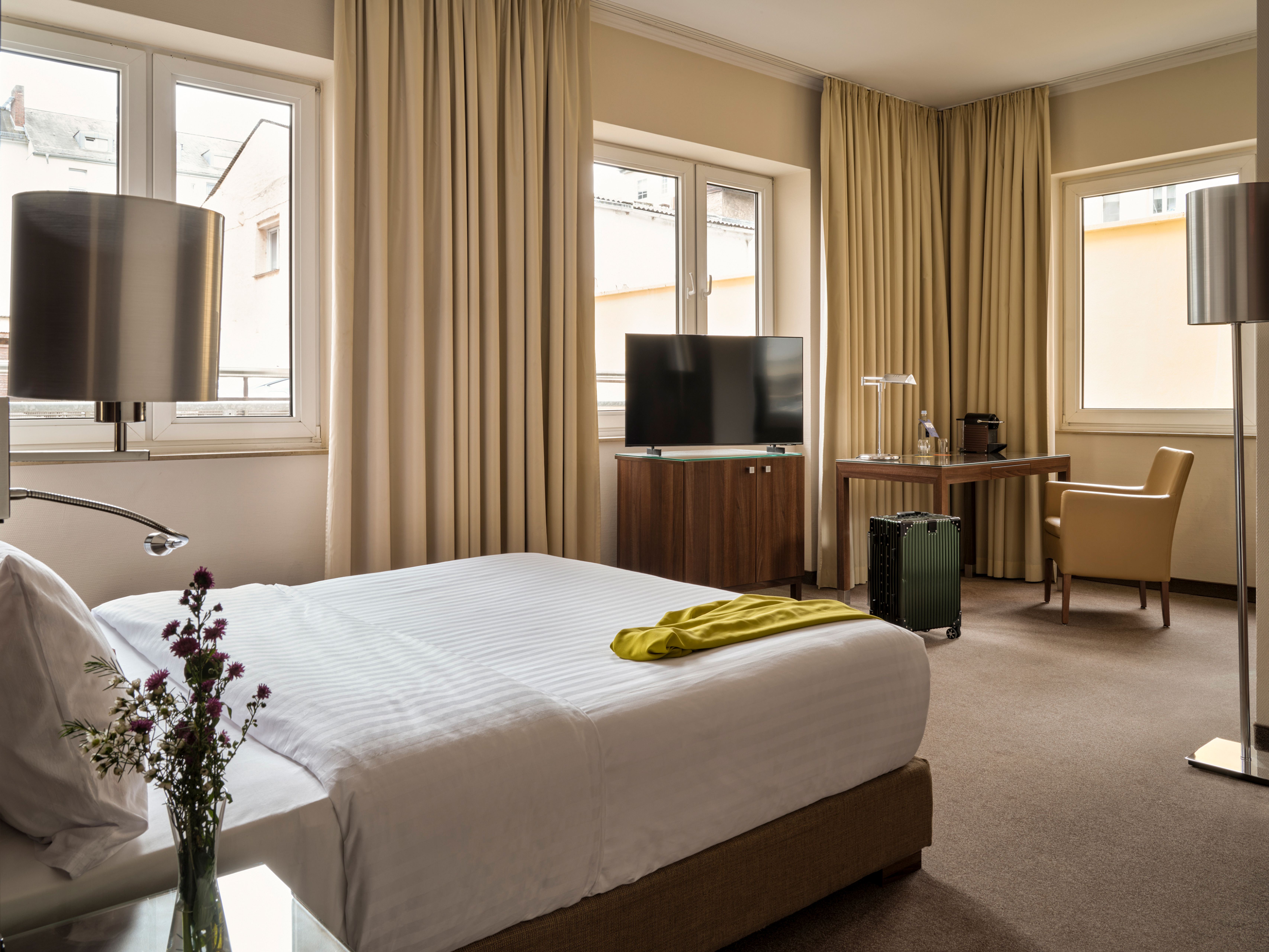 Helles Hotelzimmer mit großem Doppelbett, Fernseher, Schreibtisch und Sitzbereich. Drei Fenster bieten Tageslicht und Blick auf benachbarte Gebäude. Schlichte, warme Farben und moderne Einrichtung schaffen eine angenehme Atmosphäre. Helles Hotelzimmer mit großem Doppelbett, Fernseher, Schreibtisch und Sitzbereich. Drei Fenster bieten Tageslicht und Blick auf benachbarte Gebäude. Schlichte, warme Farben und moderne Einrichtung schaffen eine angenehme Atmosphäre.