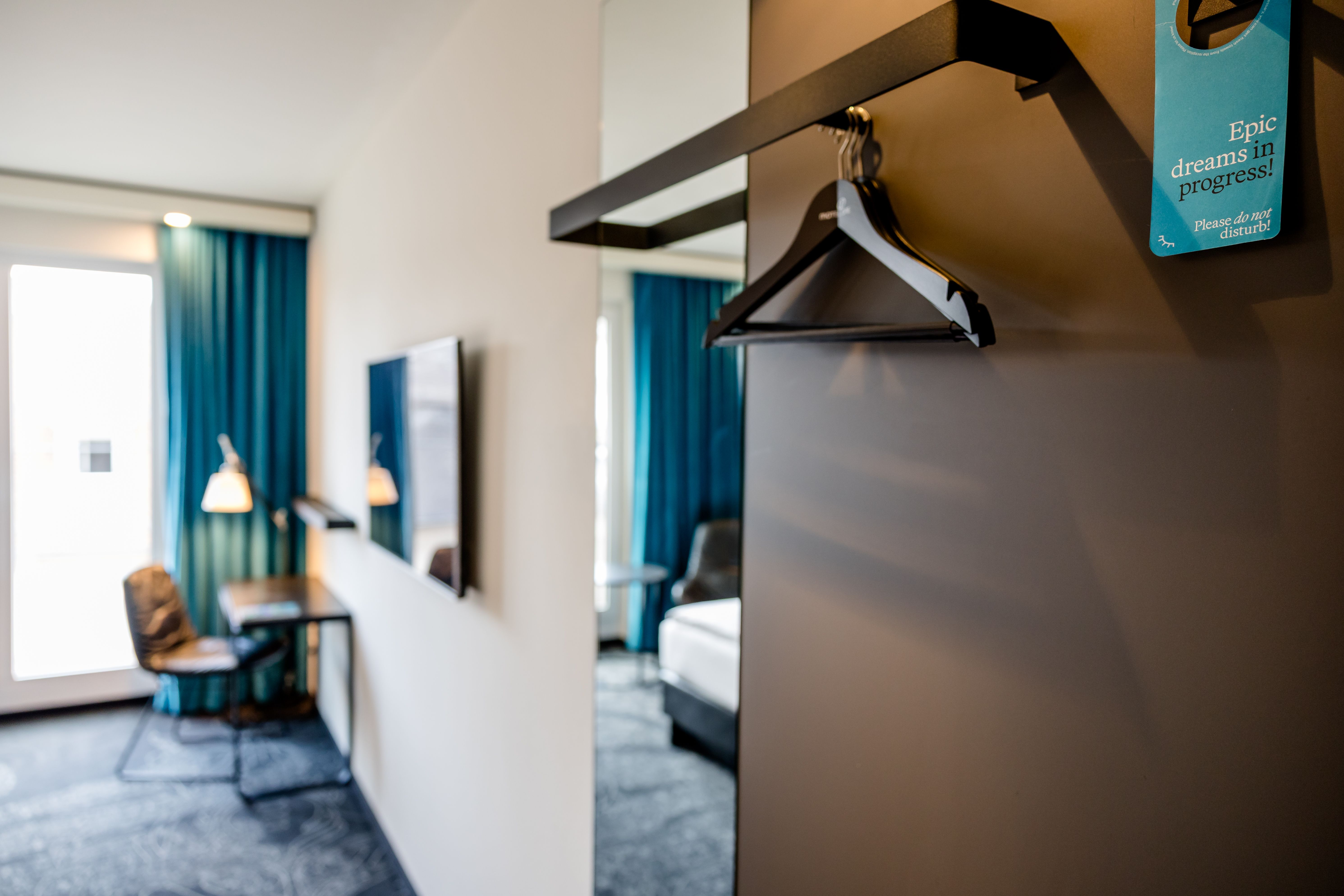Moderner Hotelzimmer-Eingangsbereich mit offenen Kleiderhaken, Blick in einen hellen Raum mit Schreibtisch, Stuhl, Fernseher und blauen Vorhängen, der Komfort und Funktionalität für Geschäftsreisende und Urlaubsgäste bietet.