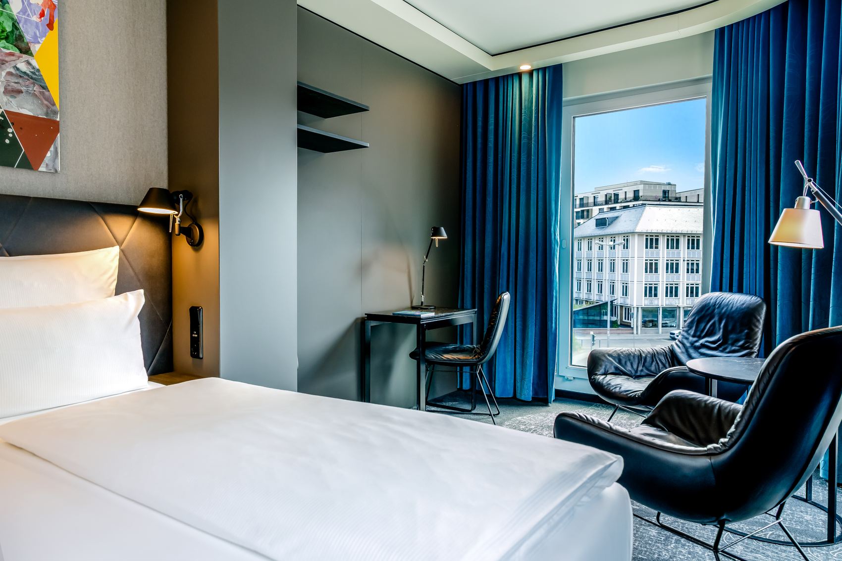 Modernes Hotelzimmer mit gemütlichem Bett, Schreibtisch, zwei komfortablen Sesselplätzen und großem Fenster mit Blick auf urbane Gebäude. Helle, freundliche Atmosphäre durch natürliche Beleuchtung.