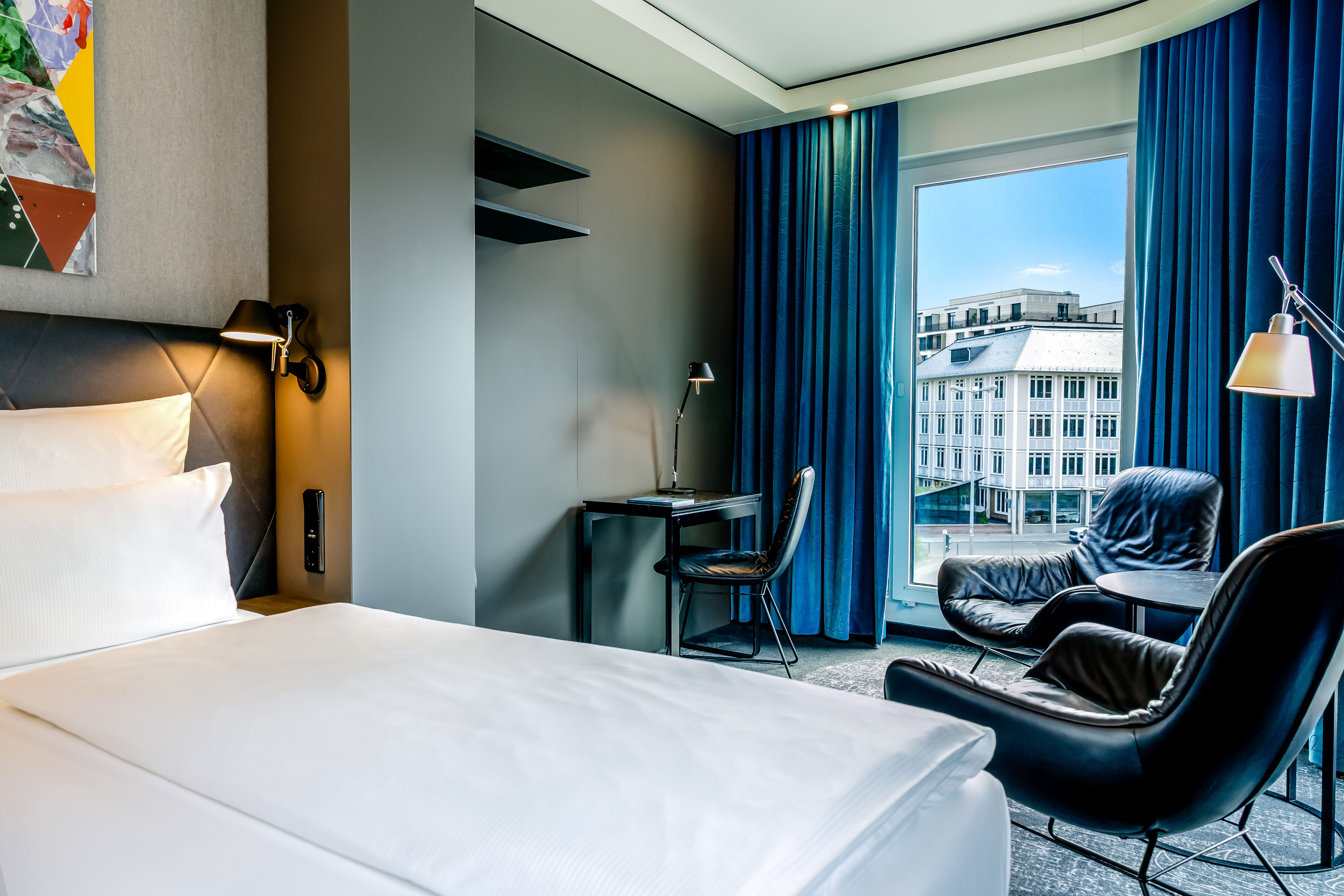 Modernes Hotelzimmer mit gemütlichem Bett, Schreibtisch, zwei komfortablen Sesselplätzen und großem Fenster mit Blick auf urbane Gebäude. Helle, freundliche Atmosphäre durch natürliche Beleuchtung. Modernes Hotelzimmer mit gemütlichem Bett, Schreibtisch, zwei komfortablen Sesselplätzen und großem Fenster mit Blick auf urbane Gebäude. Helle, freundliche Atmosphäre durch natürliche Beleuchtung.