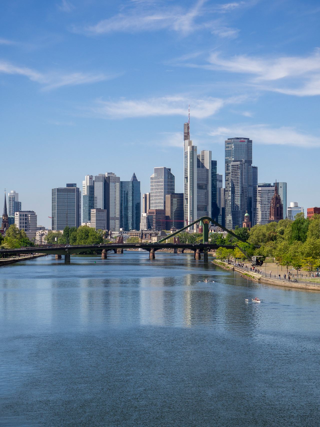 Blick auf die moderne Skyline einer Stadt mit hohen Glasgebäuden, einem Fluss im Vordergrund und einer grünen Uferpromenade bei klarem Himmel.