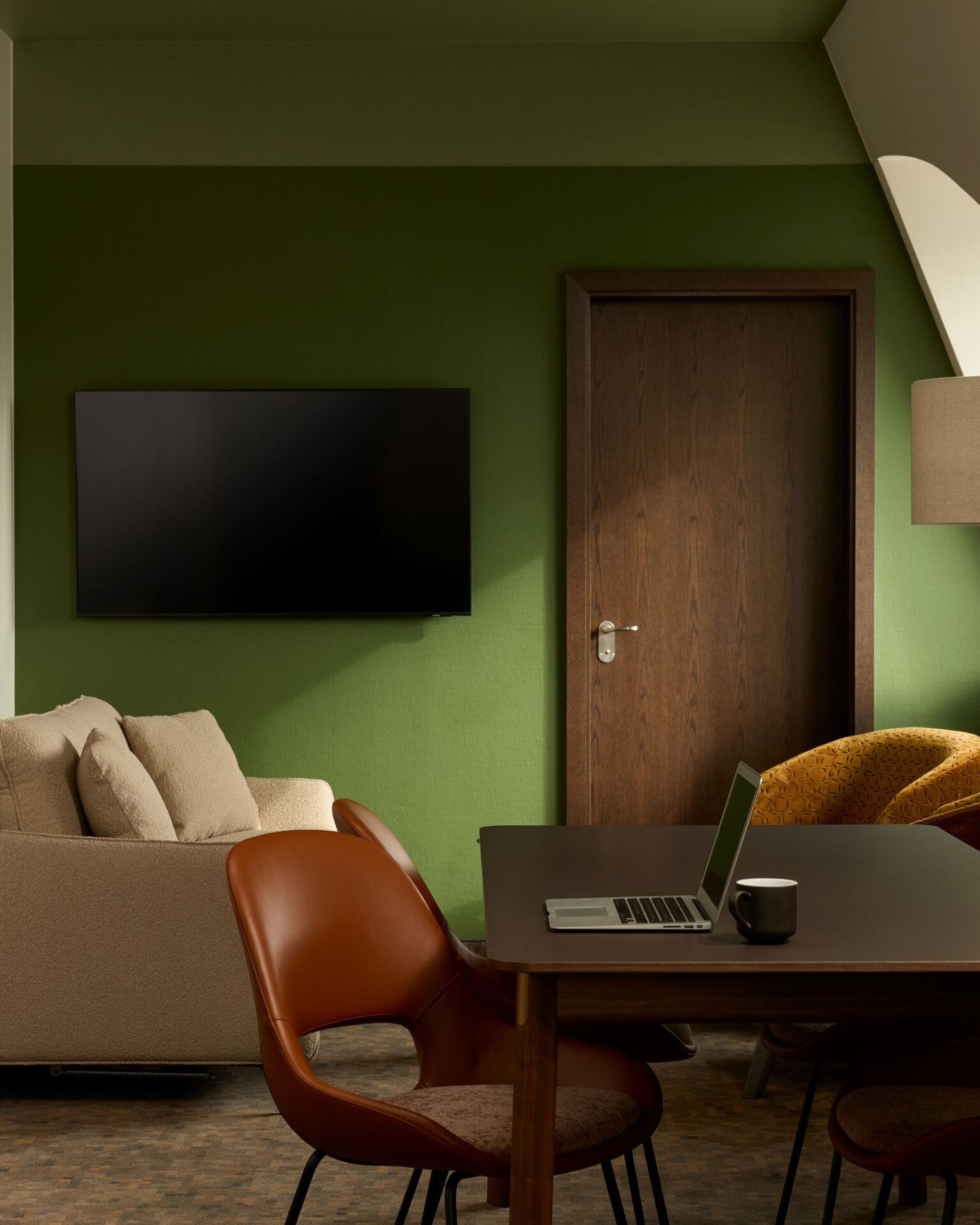 Hotel Frankfurt Metropolitan The Cloud One Zimmer mit grün gestrichener Wand, beigem Sofa, Flachbildfernseher, Holztisch mit Laptop und Kaffeetasse sowie mehreren Stühlen und Lampe.