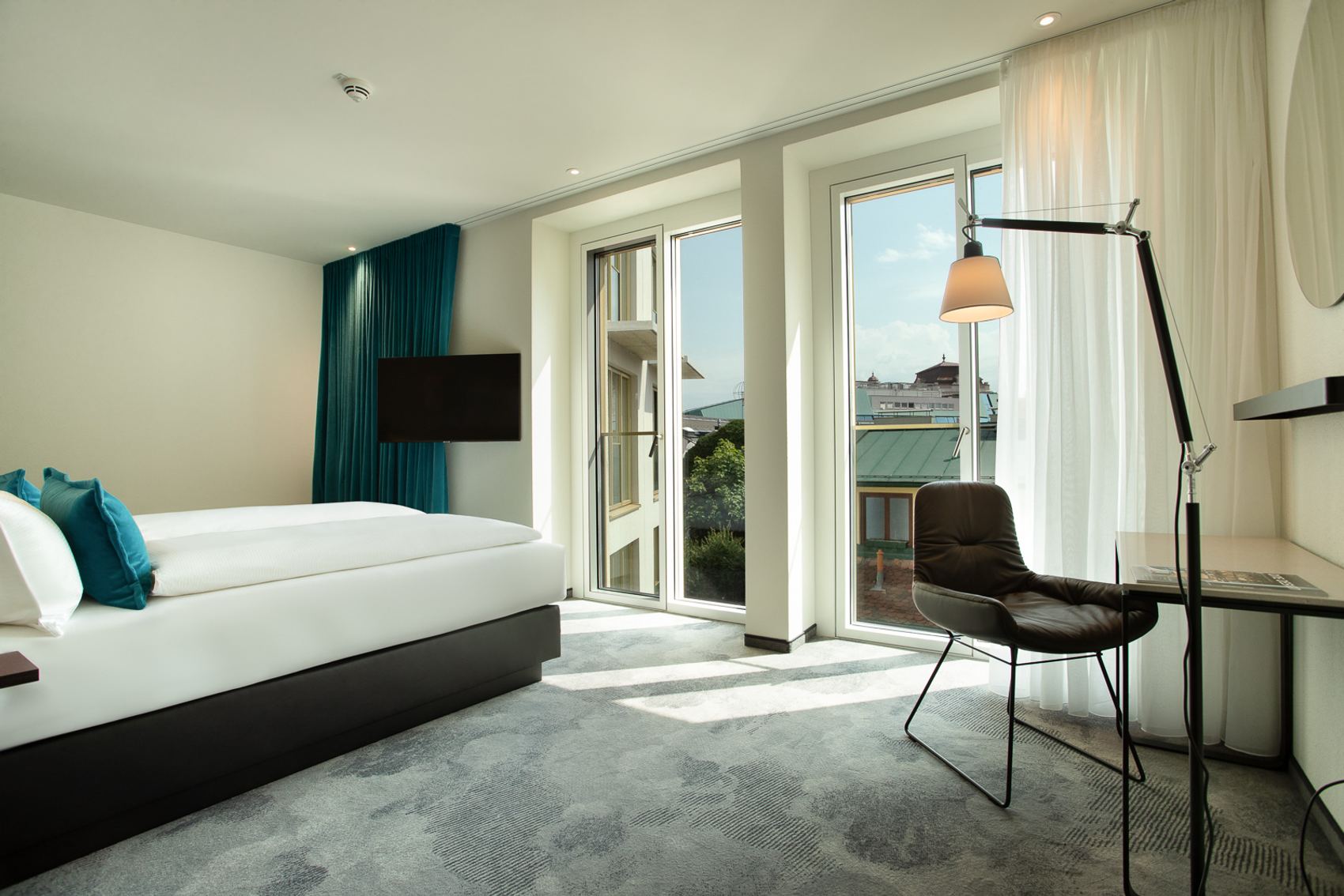 Helles Hotelzimmer mit großem Bett, bodentiefen Fenstern und Balkon, moderner Arbeitsbereich mit Stuhl und Lampe, Blick auf umliegende Dächer und grünes Dauerkleid, ruhige, einladende Atmosphäre.