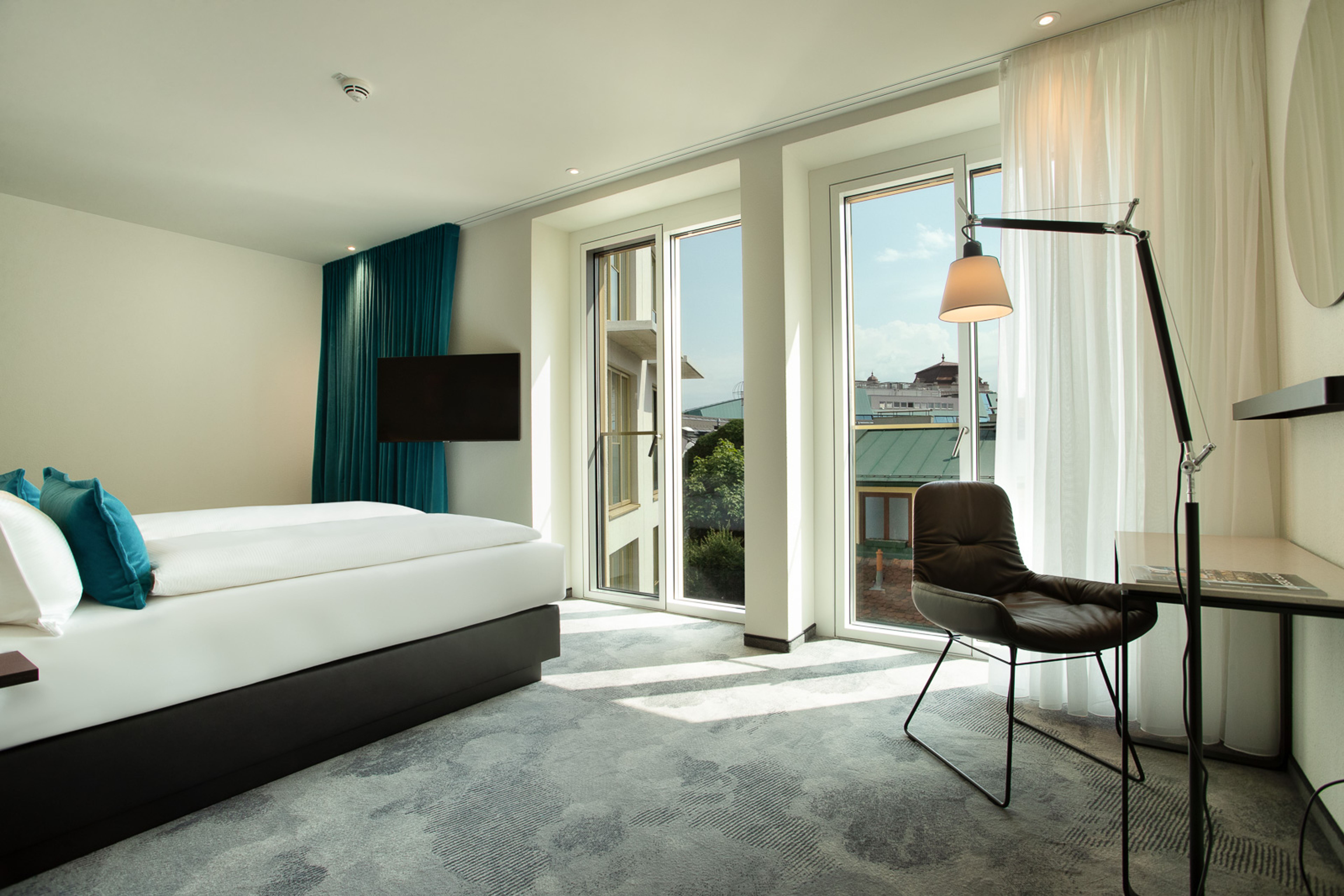 Helles Hotelzimmer mit großem Bett, bodentiefen Fenstern und Balkon, moderner Arbeitsbereich mit Stuhl und Lampe, Blick auf umliegende Dächer und grünes Dauerkleid, ruhige, einladende Atmosphäre. Helles Hotelzimmer mit großem Bett, bodentiefen Fenstern und Balkon, moderner Arbeitsbereich mit Stuhl und Lampe, Blick auf umliegende Dächer und grünes Dauerkleid, ruhige, einladende Atmosphäre.