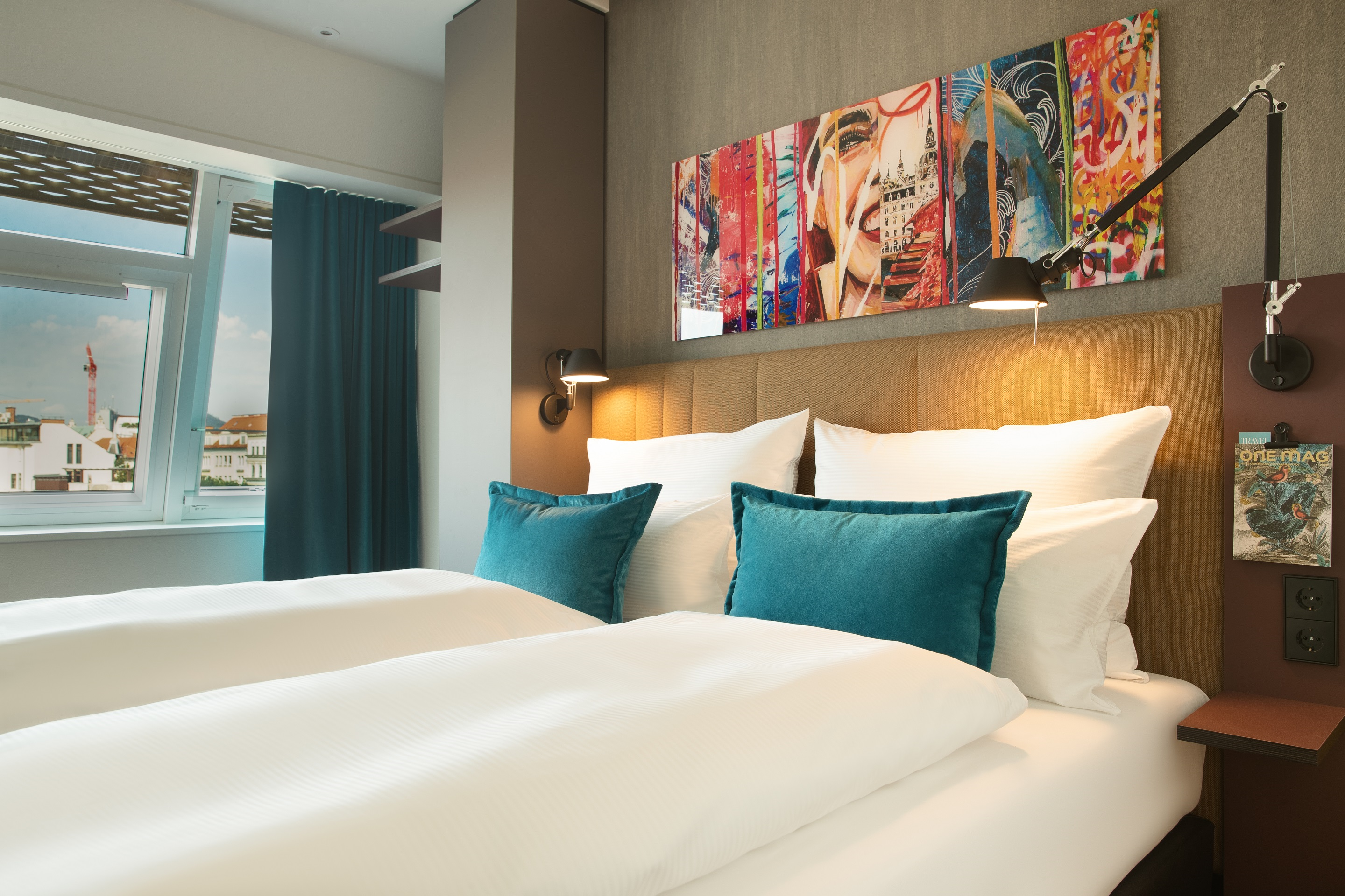 Hotel Graz Motel One großes Doppelbett mit zwei Zierkissen in türkis sowie farbenfrohes Gemälde über dem Bett