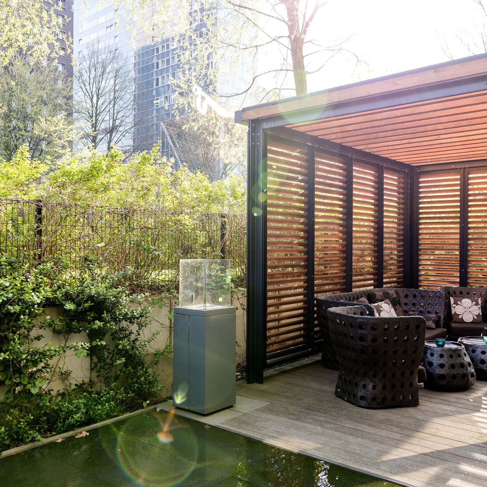 Überdachte outdoor Lounge mit schwarzem Korbmöbel-Set, Sitzkissen und Blick auf ruhigen Gartenbereich mit Wasserbecken, umgeben von Stadtgebäuden.