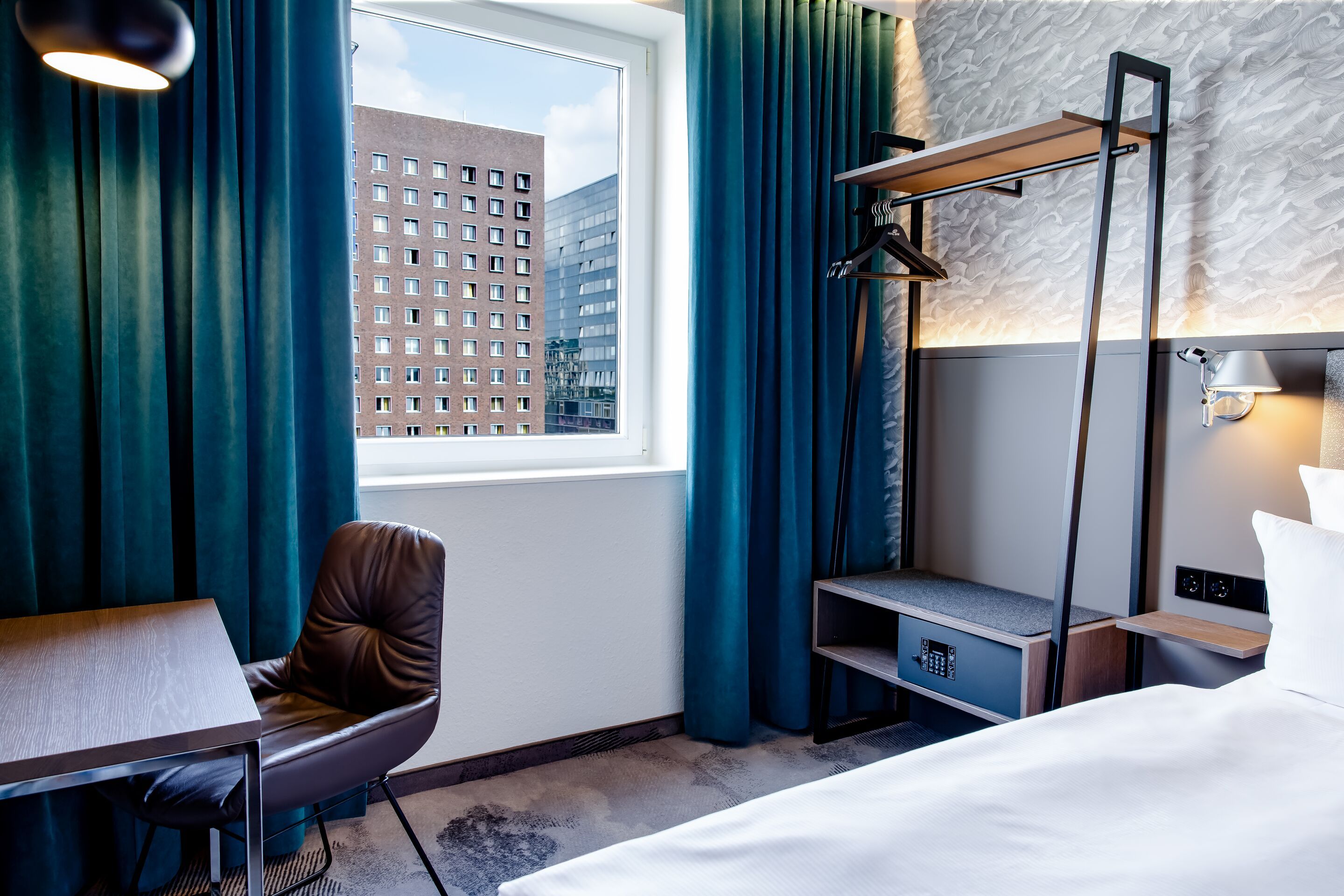 Hotel Hamburg-Alster Motel One Zimmer mit großem Fenster, Sicht auf städtische Gebäude, gemütlichem Arbeitsplatz mit Stuhl, offenem Garderobenständer und bodentiefen Vorhängen in türkis. 