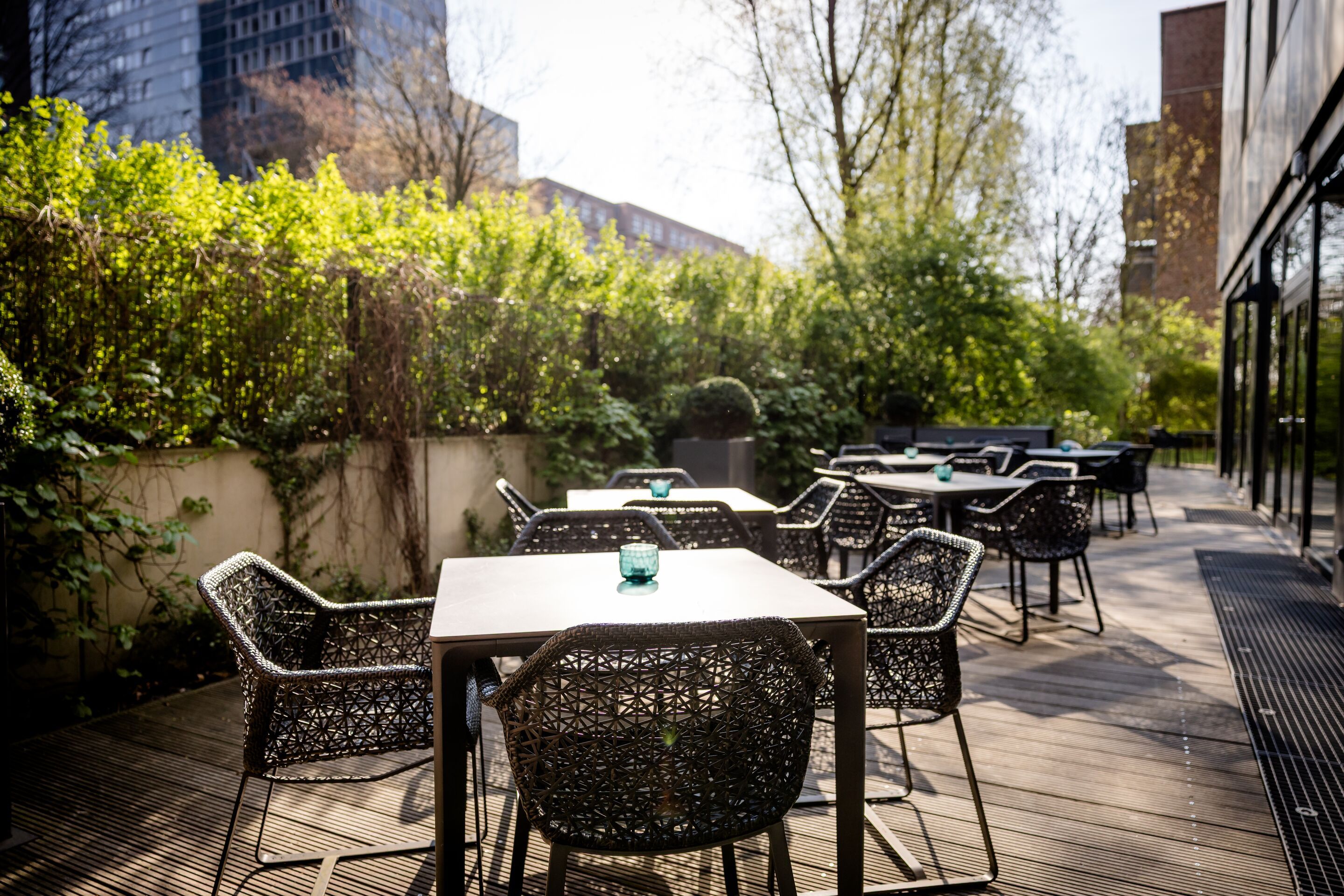 Hotel Hamburg-Alster Motel One Außenbereich mit Terrasse, mehreren Tischen und Stühlen aus geflochtenem Material, umgeben von grünen Pflanzen und Bäumen.