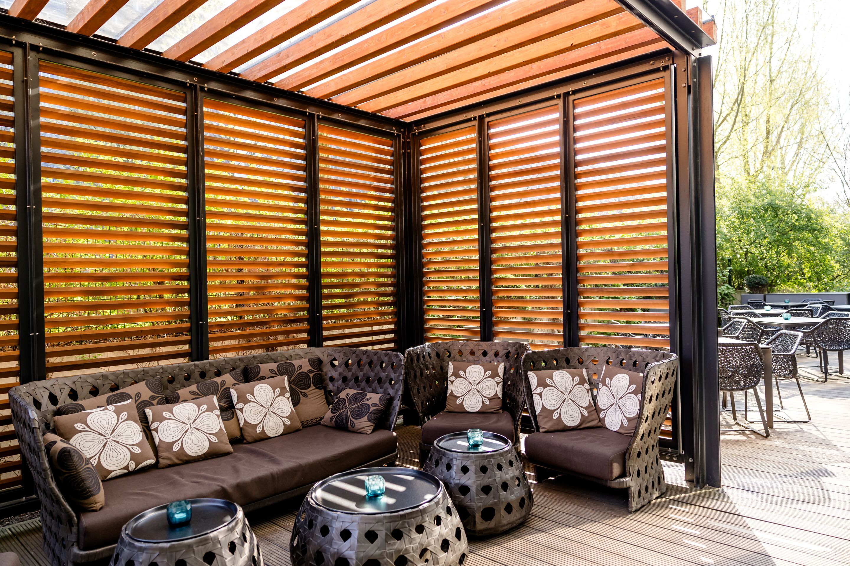 Hotel Hamburg-Alster Motel One gemütliche Lounge-Ecke mit Polstermöbeln und dekorativen Kissen unter einer Holzlamellenüberdachung, umgeben von natürlichem Licht und Blick ins Grüne.