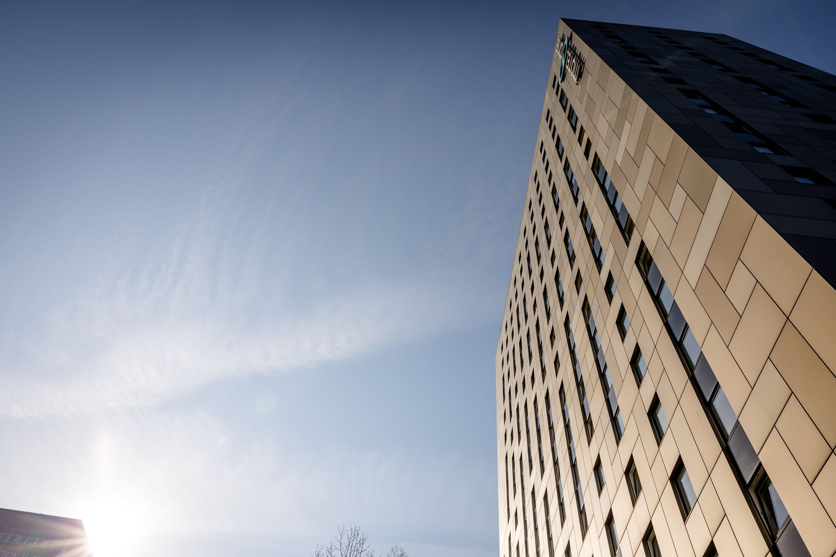 Designhotel Hamburg-Alster Motel One Hochhaus mit Fassade aus goldfarbenen und schwarzen Paneelen unter klarem, blauem Himmel, aufgenommen aus der Froschperspektive.