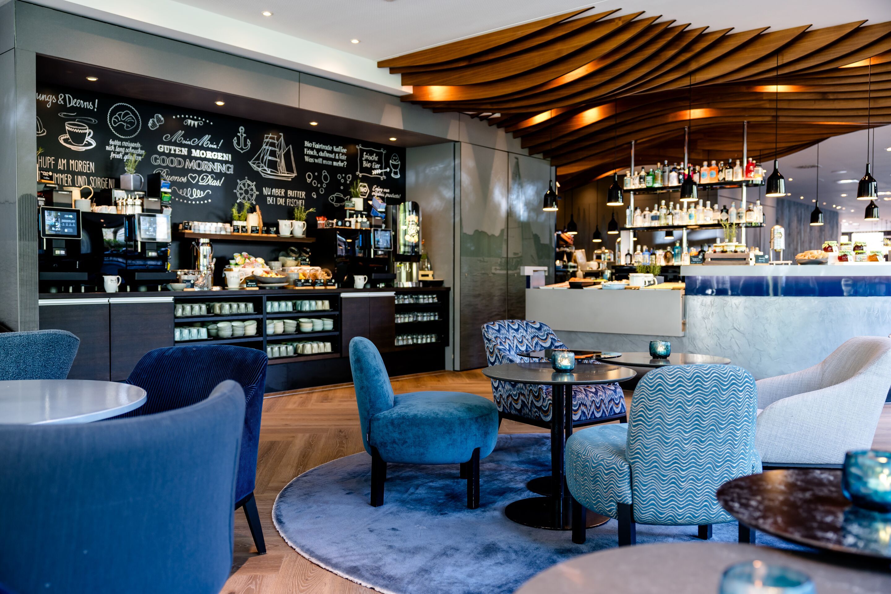 Hotel Hamburg-Alster Motel One Lounge und Frühstücksbereich mit bequemen Sesseln und stilvoller Beleuchtung, im Hintergrund Buffet mit Auswahl an Getränken und Kaffeemaschine.