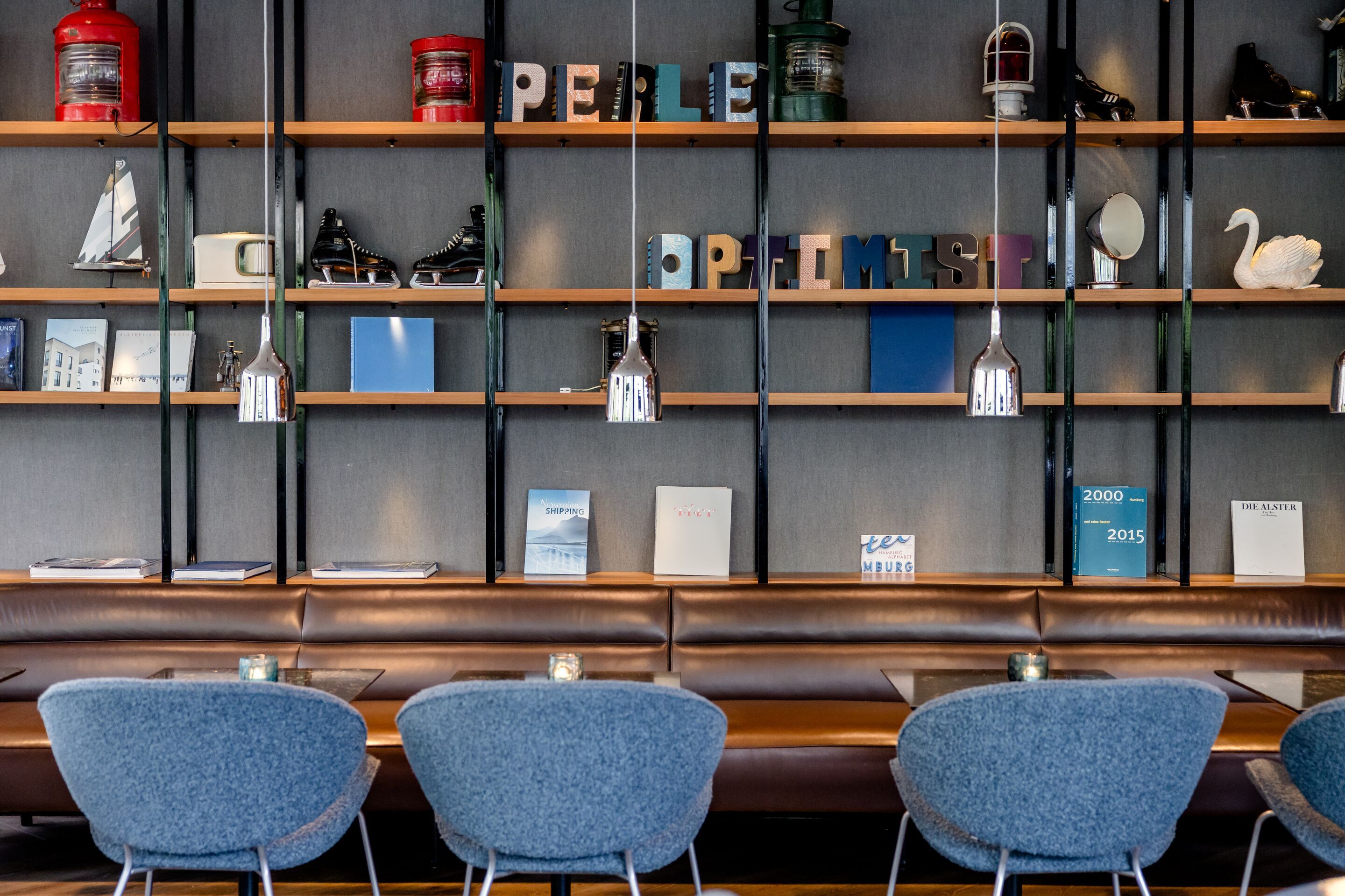 Hotel Hamburg-Alster Motel One Lounge mit blauen Sesseln, braunem Ledersofa und Wandregal, das dekorative Elemente und Buchstaben zeigt. 
