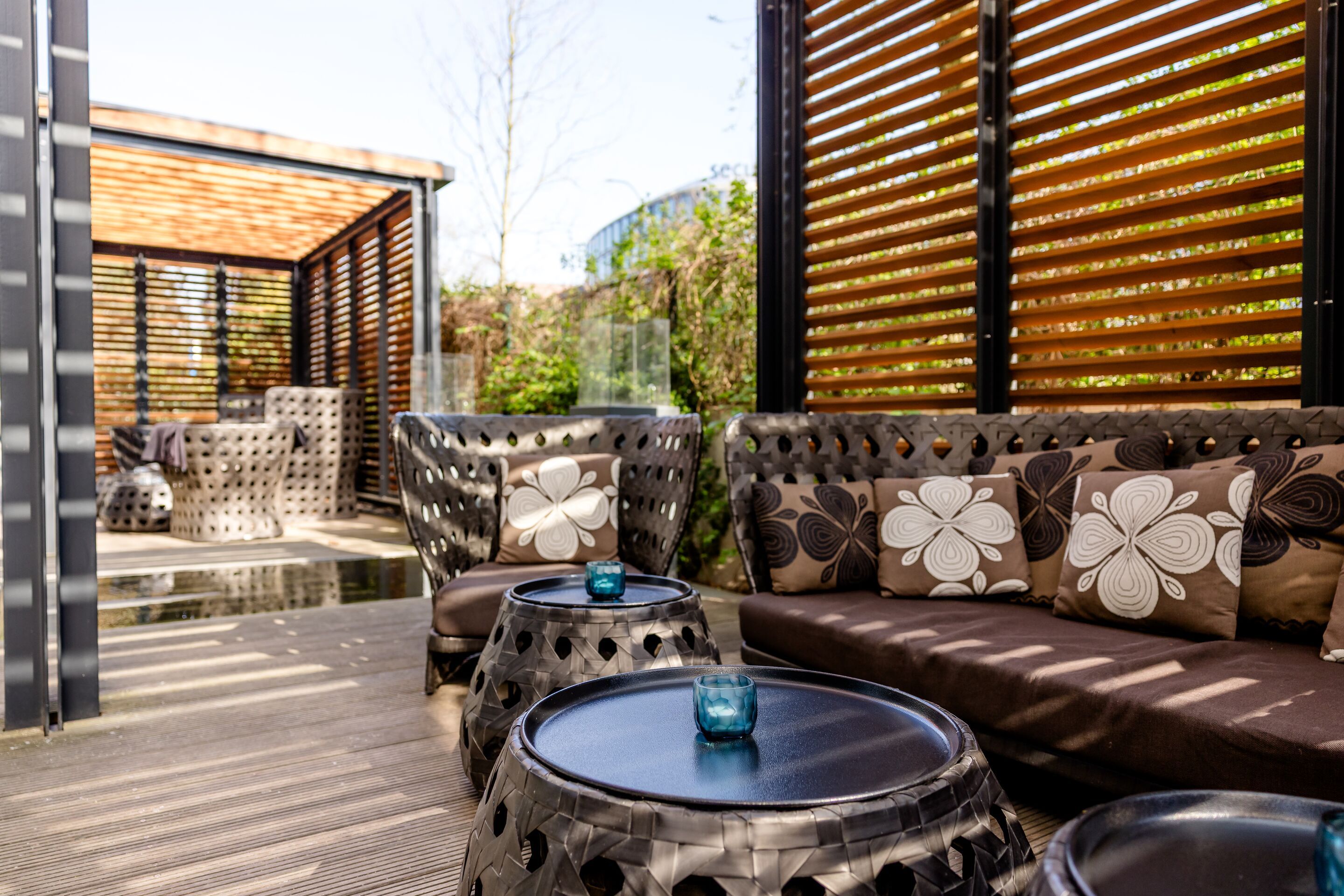 Hotel Hamburg-Alster Motel One Außensitzecke mit gepolsterten Sofas und Sesseln, dekoriert mit blumigen Kissen, umgeben von Holzlamellen für Privatsphäre.