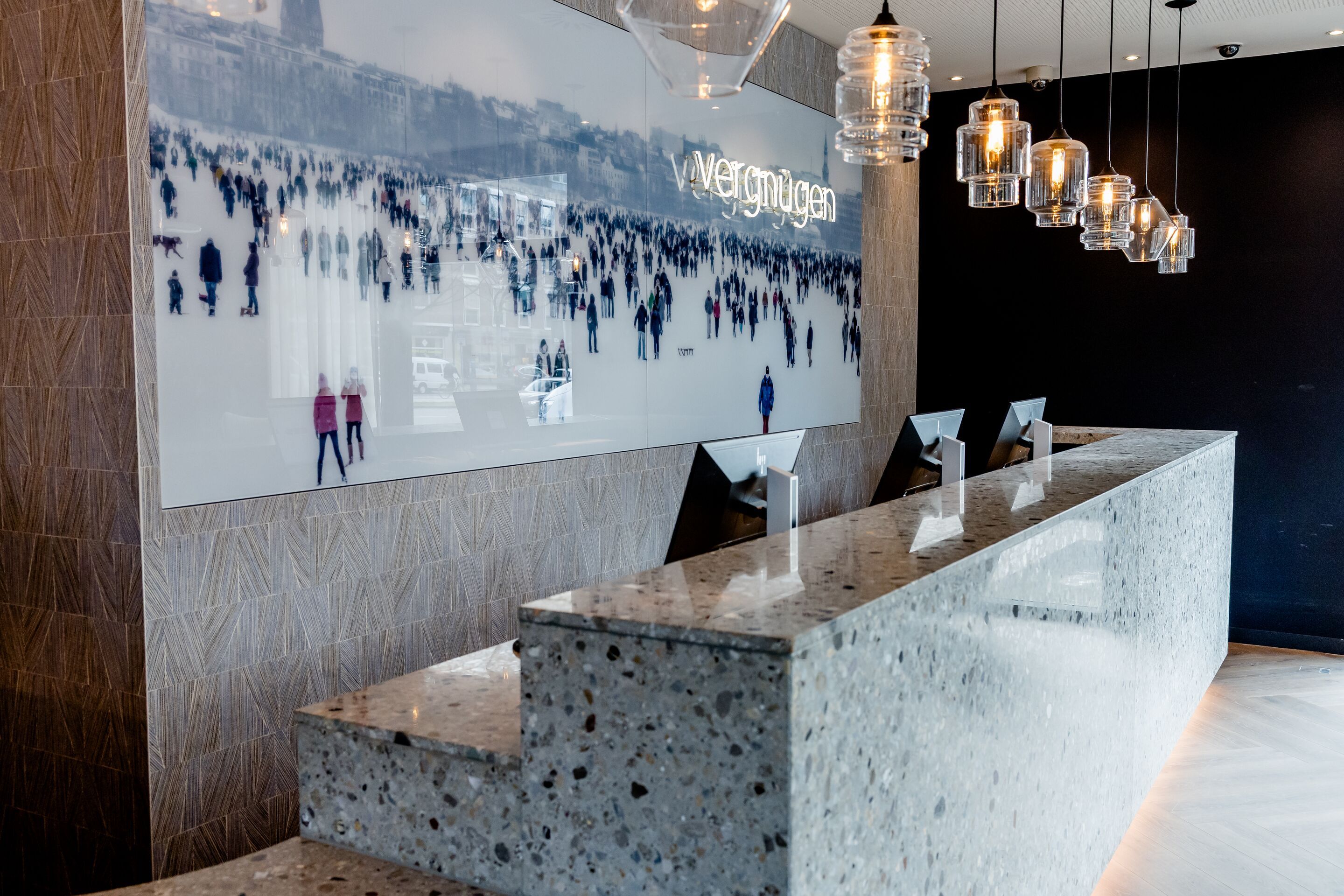 Hotel Hamburg-Alster Motel One Rezeption mit langer, grauer Stein-Theke, drehbaren Monitoren und Pendelleuchten. An der Wand hängt ein großes Bild mit der Aufschrift „Vergnügen“, das eine Menschenmenge auf einer Eisfläche zeigt. 