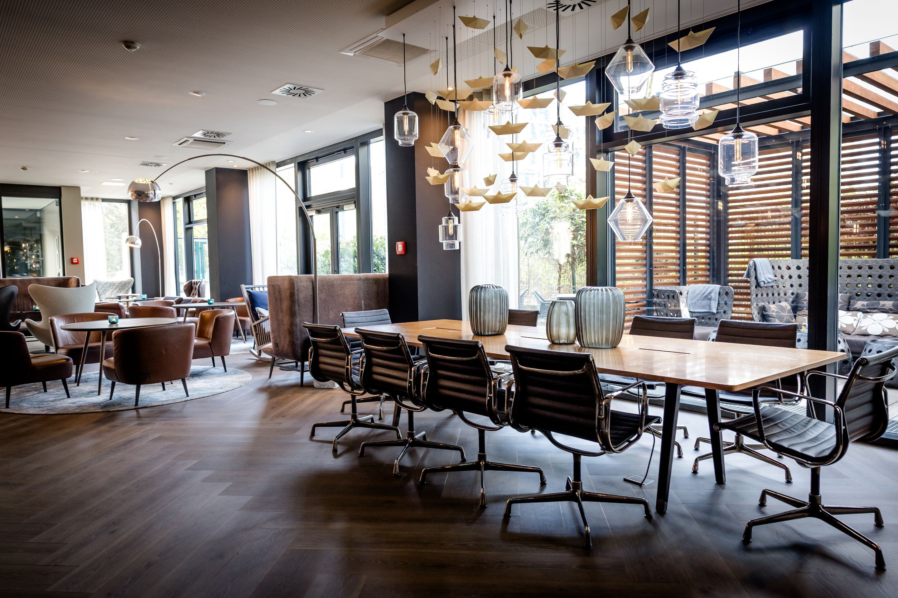 Designhotel Hamburg-Alster Motel One Lounge mit großem Holztisch, Stühlen und dekorativer Hängeleuchte. Große Fenster bieten Blick auf eine Terrasse mit Lounge-Möbeln.