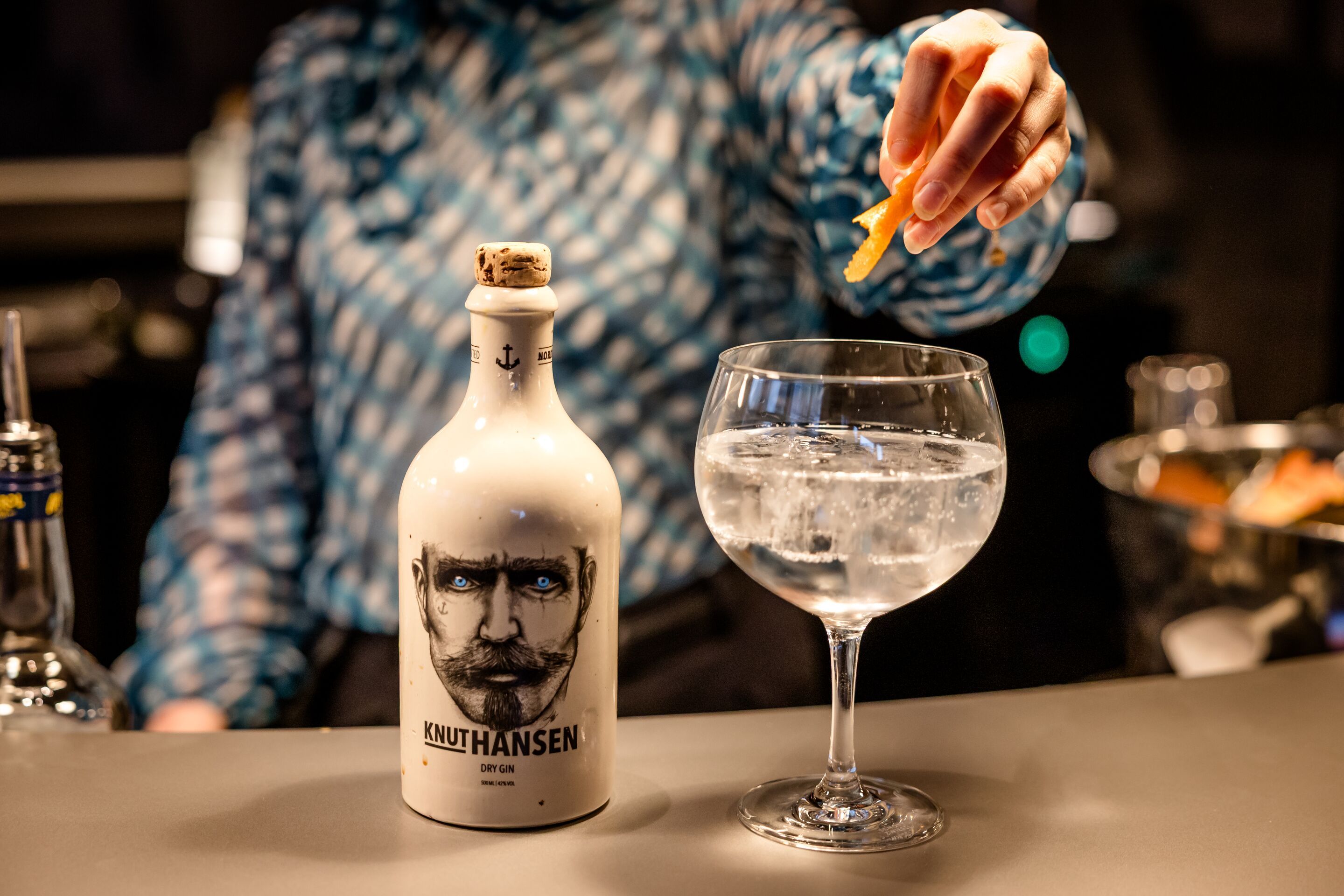 Hotel Hamburg-Alster Motel One Nahaufnahme Bar mit Barkeeperin, die einen Drink zubereitet, daneben eine weiße Flasche Gin mit Gesichtsmotiv und dem Schriftzug „Knut Hansen“.