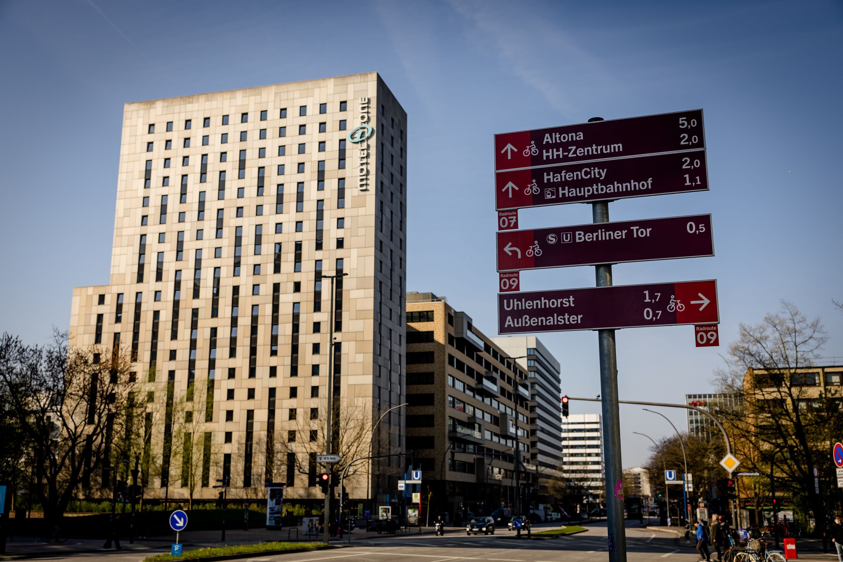 Hotel Hamburg-Alster Motel One Hotelgebäude vor klarem Himmel an einer Stadtstraße mit einem Wegweiser für Radfahrer, der Strecken und Entfernungen zu verschiedenen Zielen in Hamburg anzeigt.