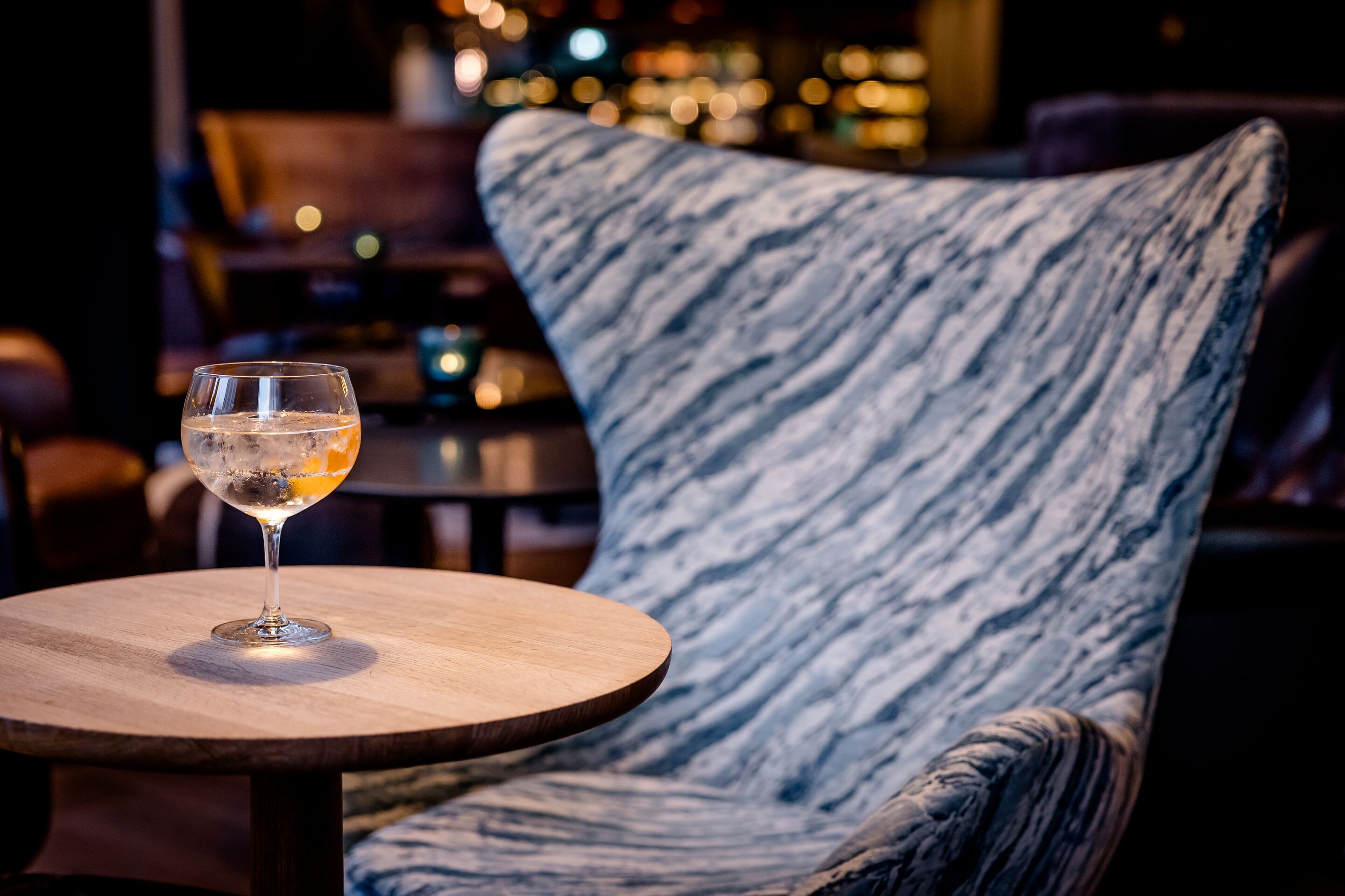 Hotel Hamburg-Alster Motel One bequemer Egg Chair Sessel neben einem kleinen Holztisch mit einem Getränk in einem eleganten Glas, in einer warm und gemütlich beleuchteten Lounge-Atmosphäre.