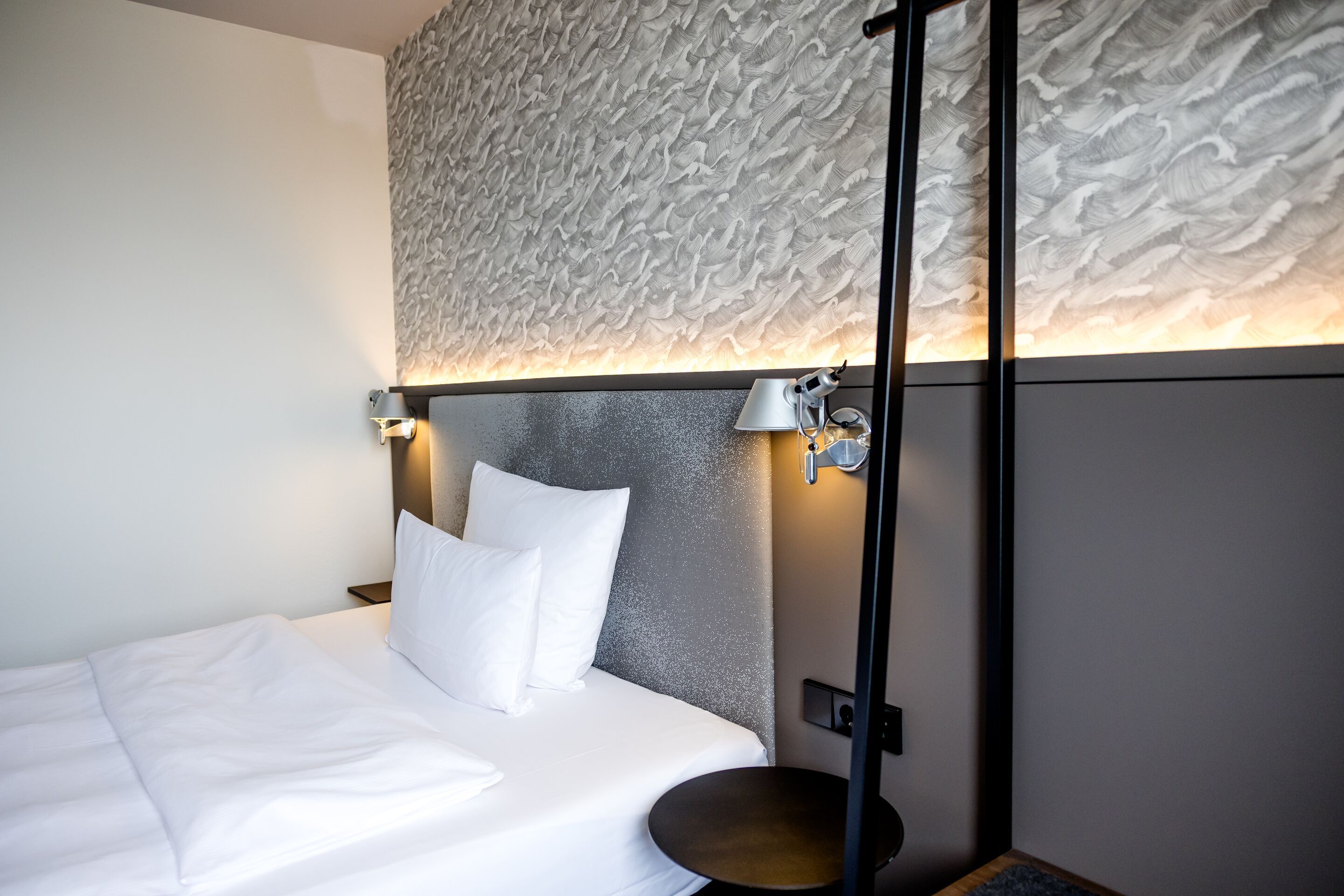 Hotel Hamburg-Alster Motel One helles Zimmer mit gemütlichem Einzelbett, weißen Kissen sowie weiße Decke und indirekte Beleuchtung.