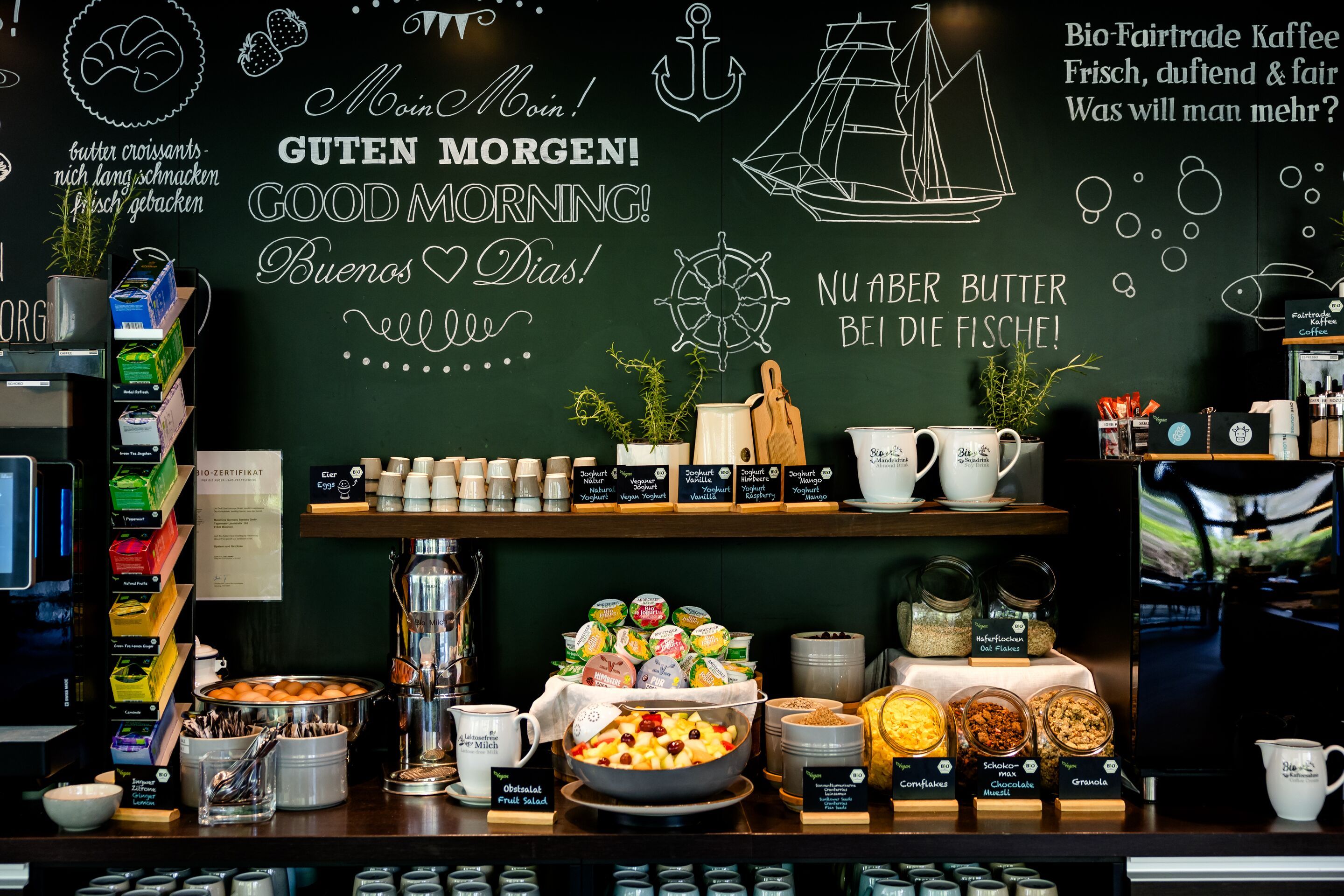 Hotel Hamburg-Alster Motel One Buffet mit frischem Frühstück, darunter Obstsalat, Gebäck, Müsli und Bio-Fairtrade-Kaffee, vor einer dekorativen Wand mit handgeschriebenen Morgengrüßen und nautischen Motiven.