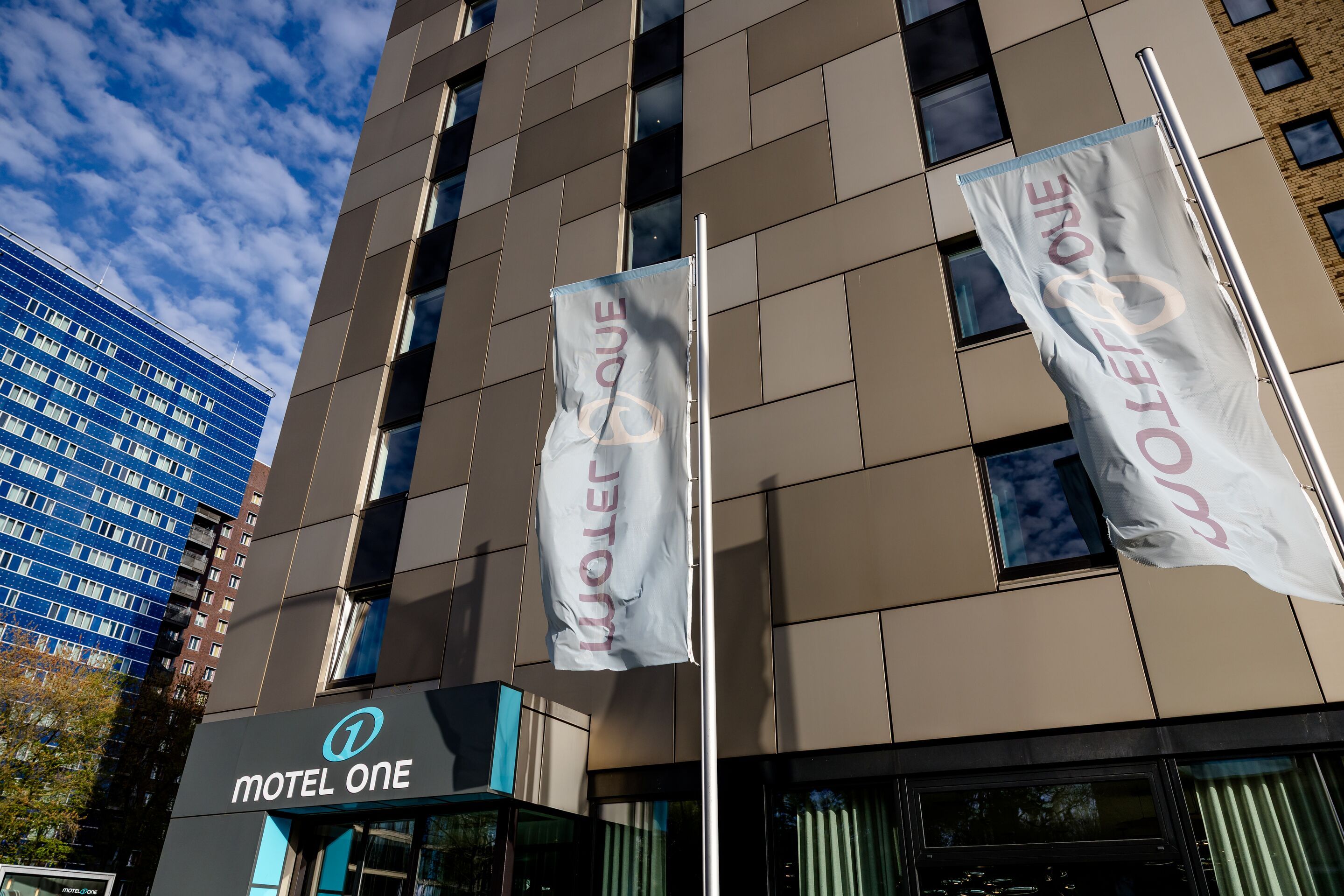 Hotel Hamburg Alster Motel One Fassade des Motel One Hotels mit zwei wehenden Fahnen und blauem Himmel im Hintergrund, sichtbarer Hoteleingang mit Logo.