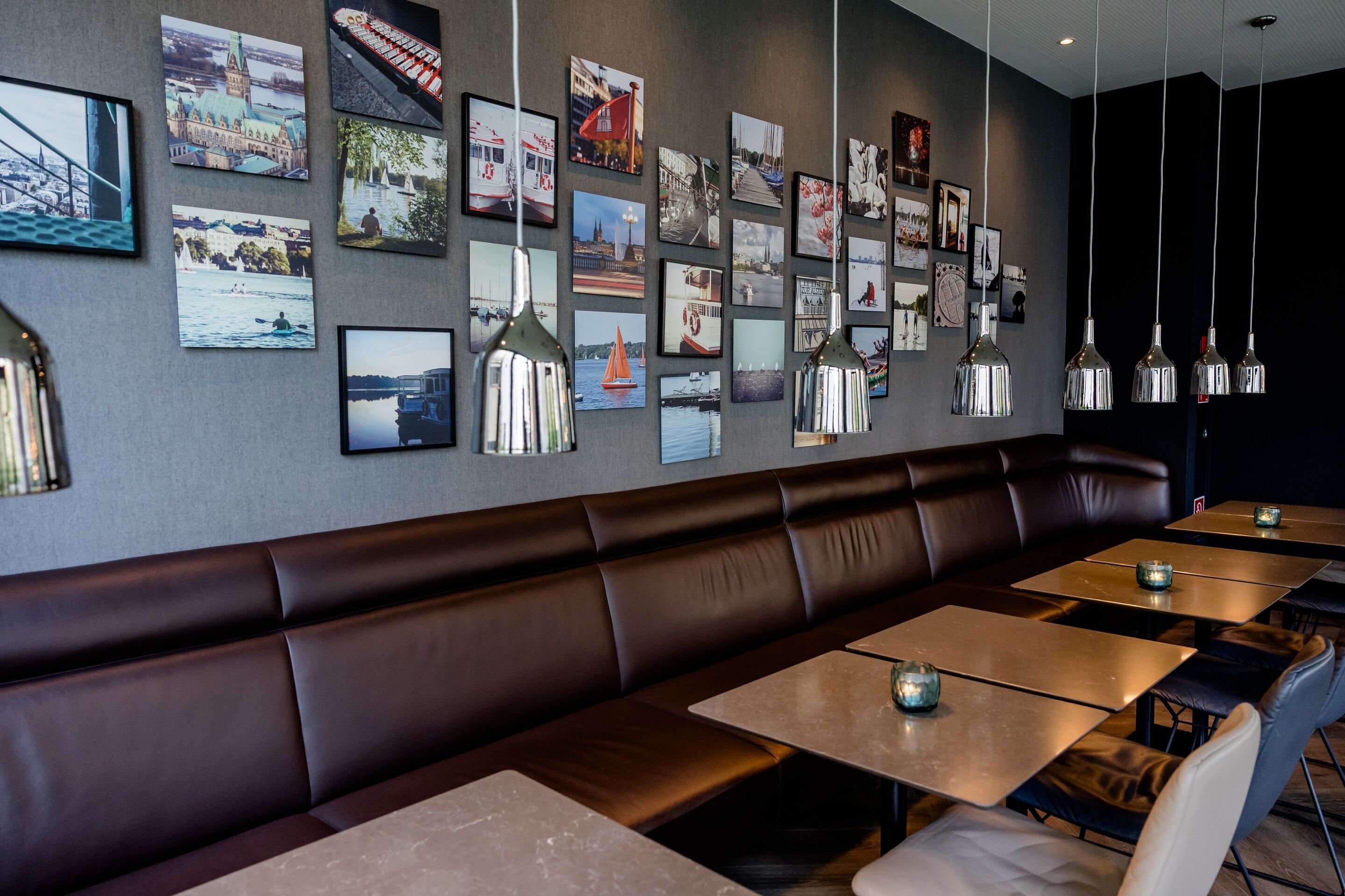 Hotel Hamburg Alster Motel One Sitzbereich mit braunen Lederbänken, Tischen und Stühlen in einem stilvollen Raum, an der Wand mehrere gerahmte Fotos und Hängeleuchten für angenehme Beleuchtung.