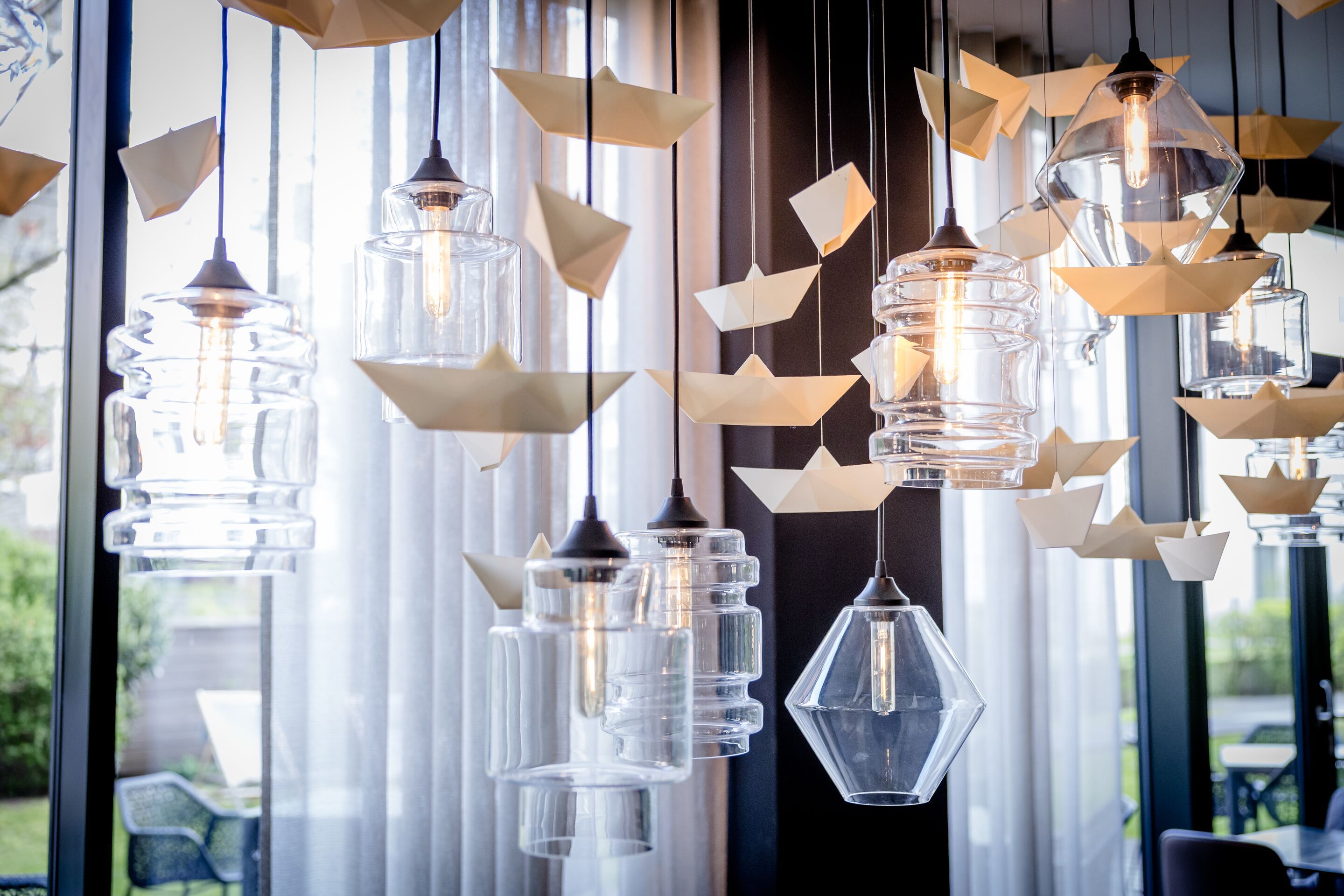 Designhotel Hamburg-Alster Motel One hängende Glaslampen und zahlreiche filigrane weiße Papierboote als dekoratives Mobile vor großen Fenstern mit lichtdurchlässigen Vorhängen.
