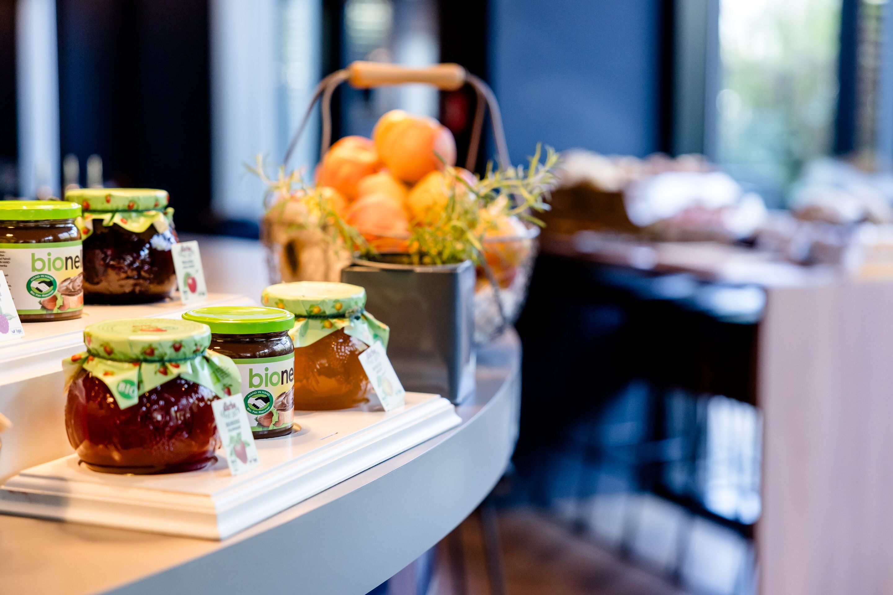 Hotel Hamburg-Alster Motel One Frühstücksbereich mit verschiedenen Bio-Marmeladen in Gläsern auf einem weißen Display, im Hintergrund ein Korb mit frischen Orangen. 