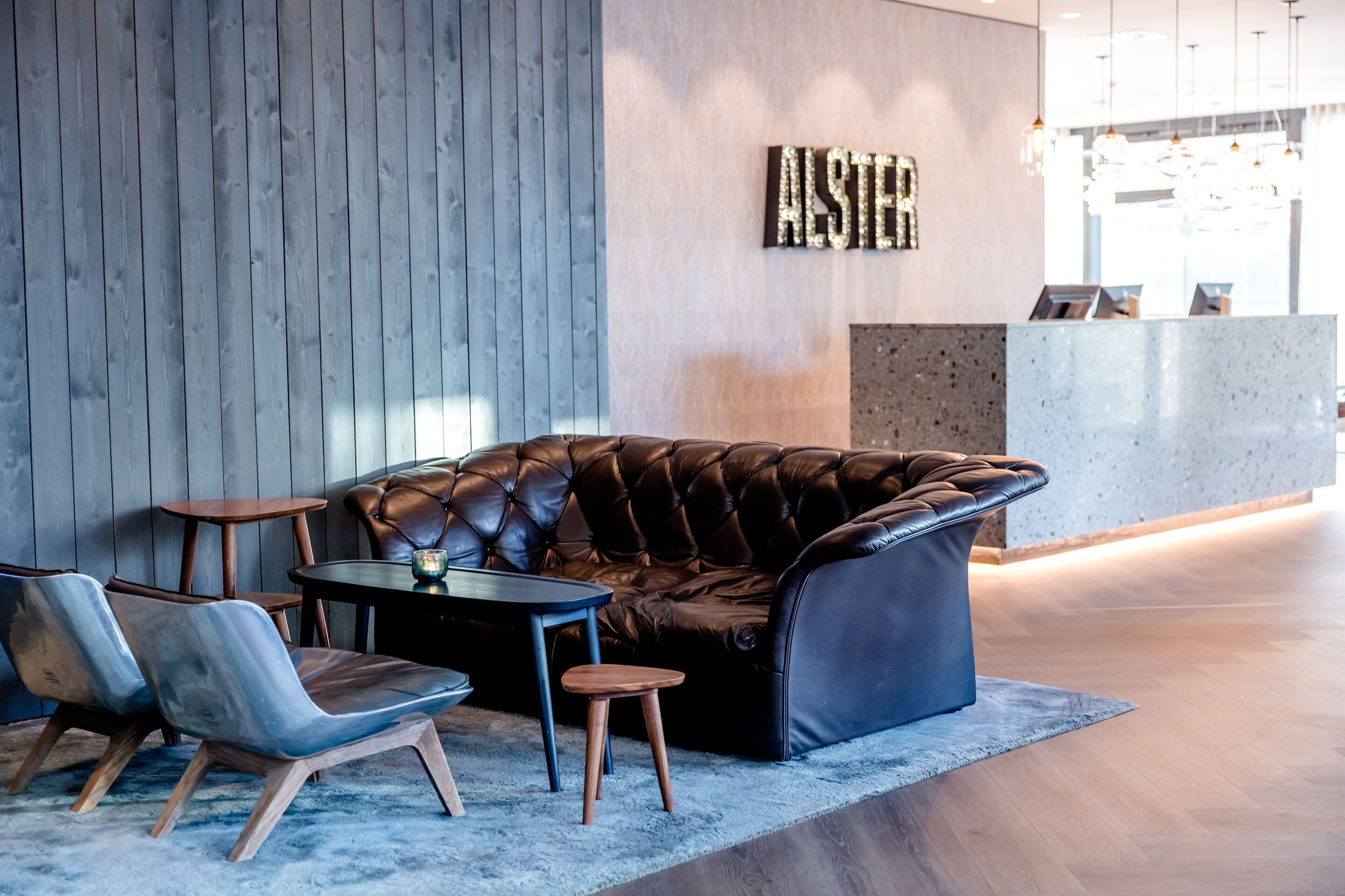 Hotel Hamburg-Alster Motel One Eingangsbereich mit bequemem schwarzen Sofa, zwei grauen Sesseln und kleiner Holztischgruppe auf Teppich, helles Ambiente mit stilvollem Empfangstresen im Hintergrund.