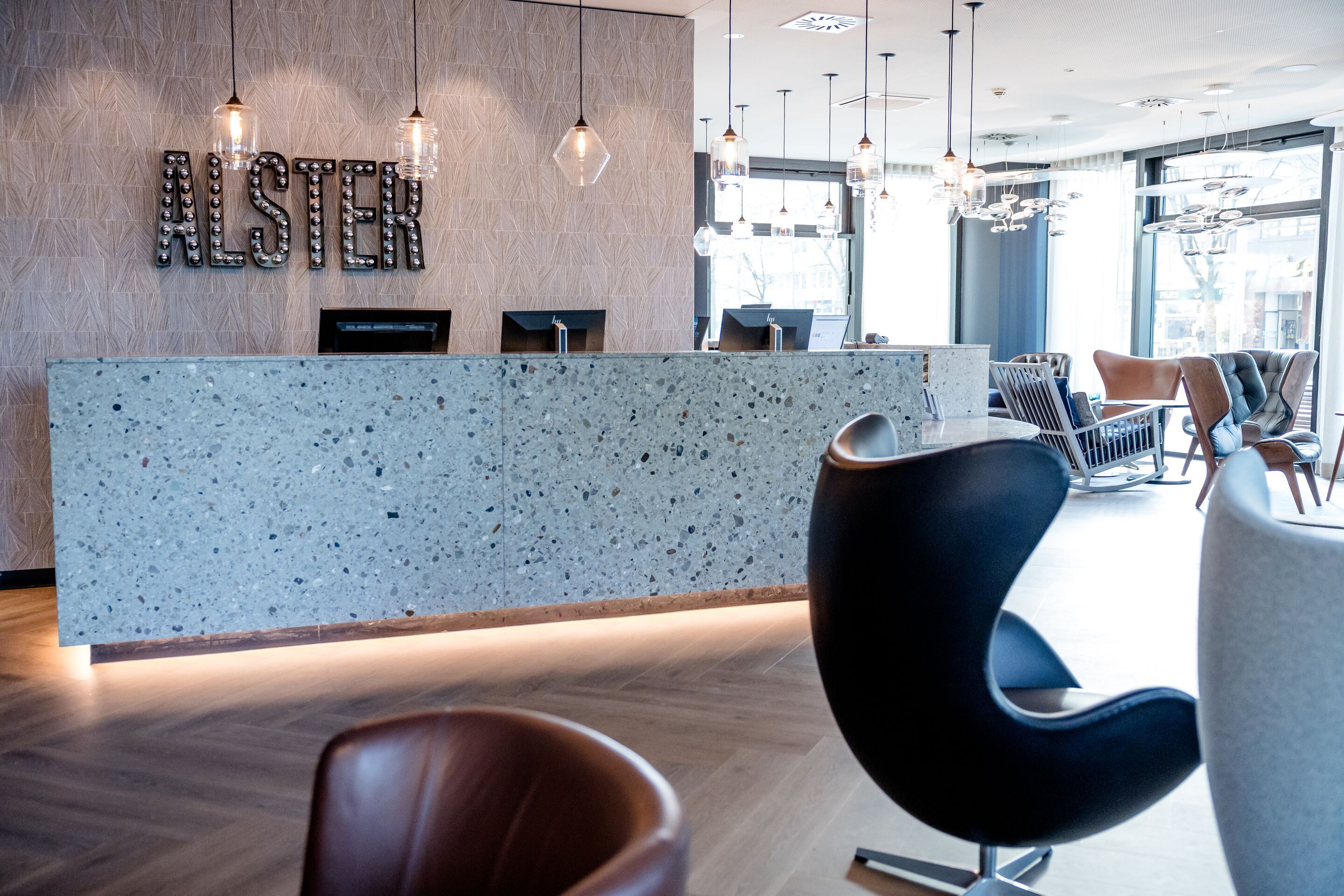 Hotel Hamburg-Alster Motel One Lounge mit hellen Holzböden, stilvoller Rezeption aus hellem Stein, bequemen Designstühlen und großer Fensterfront, die viel Tageslicht hereinlässt.