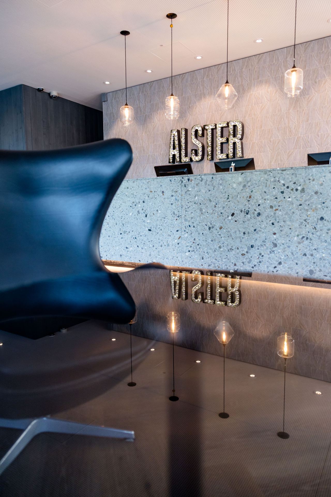 Hotel Hamburg-Alster Motel One Rezeption mit stilvoller Empfangstheke aus Stein, beleuchtet von hängenden Pendelleuchten, daneben eine elegante schwarze Lounge-Chair. Schriftzug „ALSTER“ an der Wand betont Standort und Ambiente.
