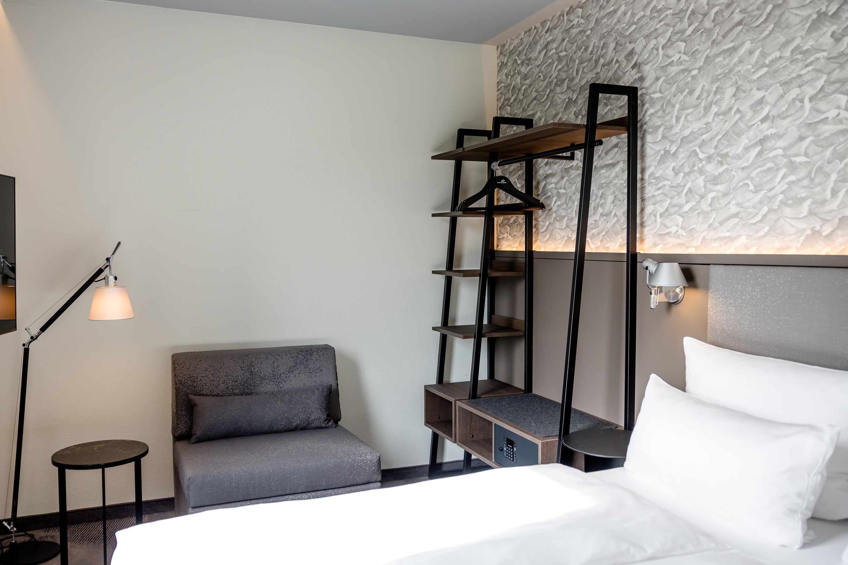 Hotel Hamburg-Alster Motel One Zimmer mit Bett und weißer Bettwäsche, grauem Sessel, offenem Garderobenregal und indirekter Beleuchtung.