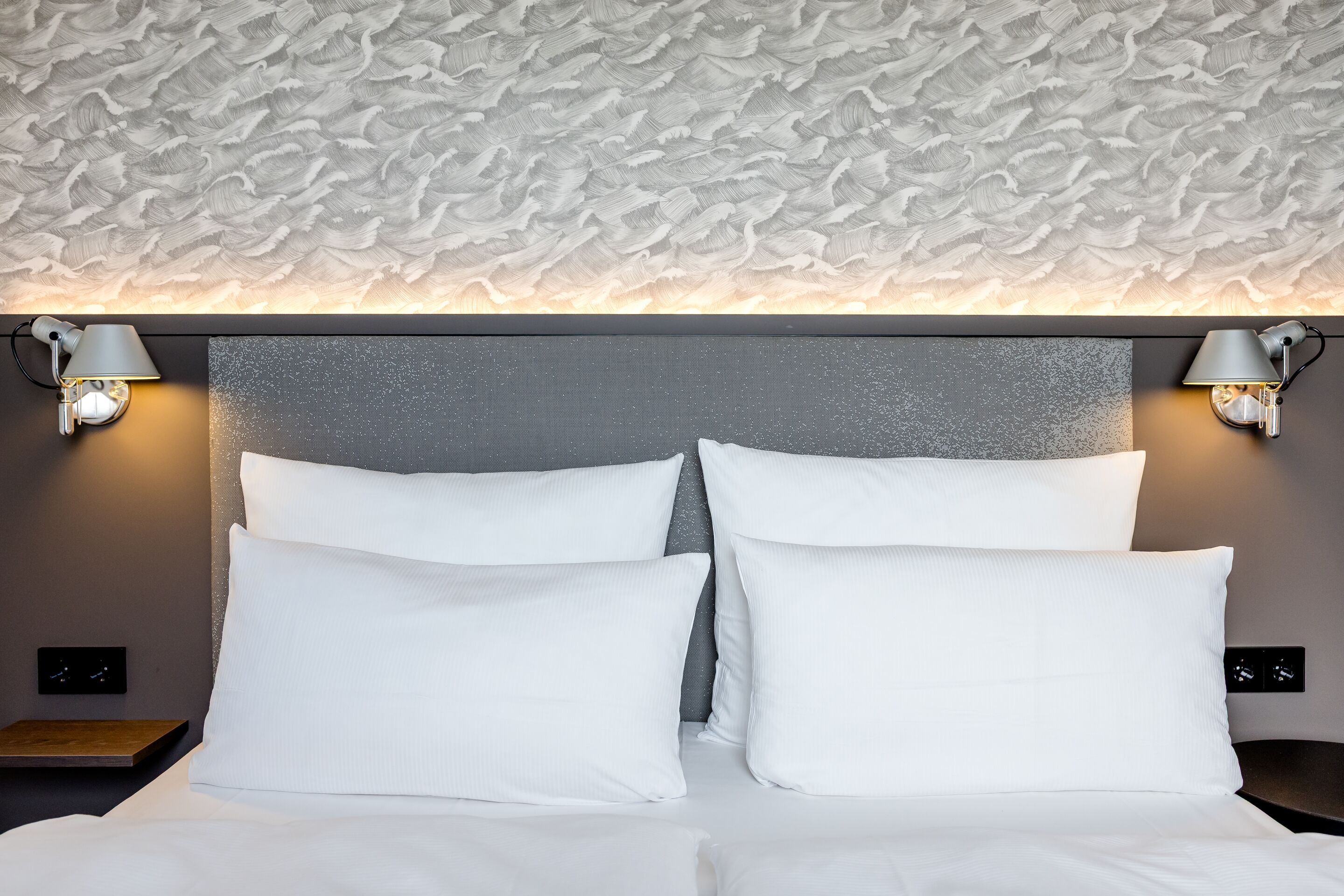 Hotel Hamburg-Alster Motel One Zimmer mit Blick auf Kopfteil eines  Bettes mit weißer Bettwäsche und mehreren Kissen sowie weiches Polsterkopfteil und warme Wandbeleuchtung.