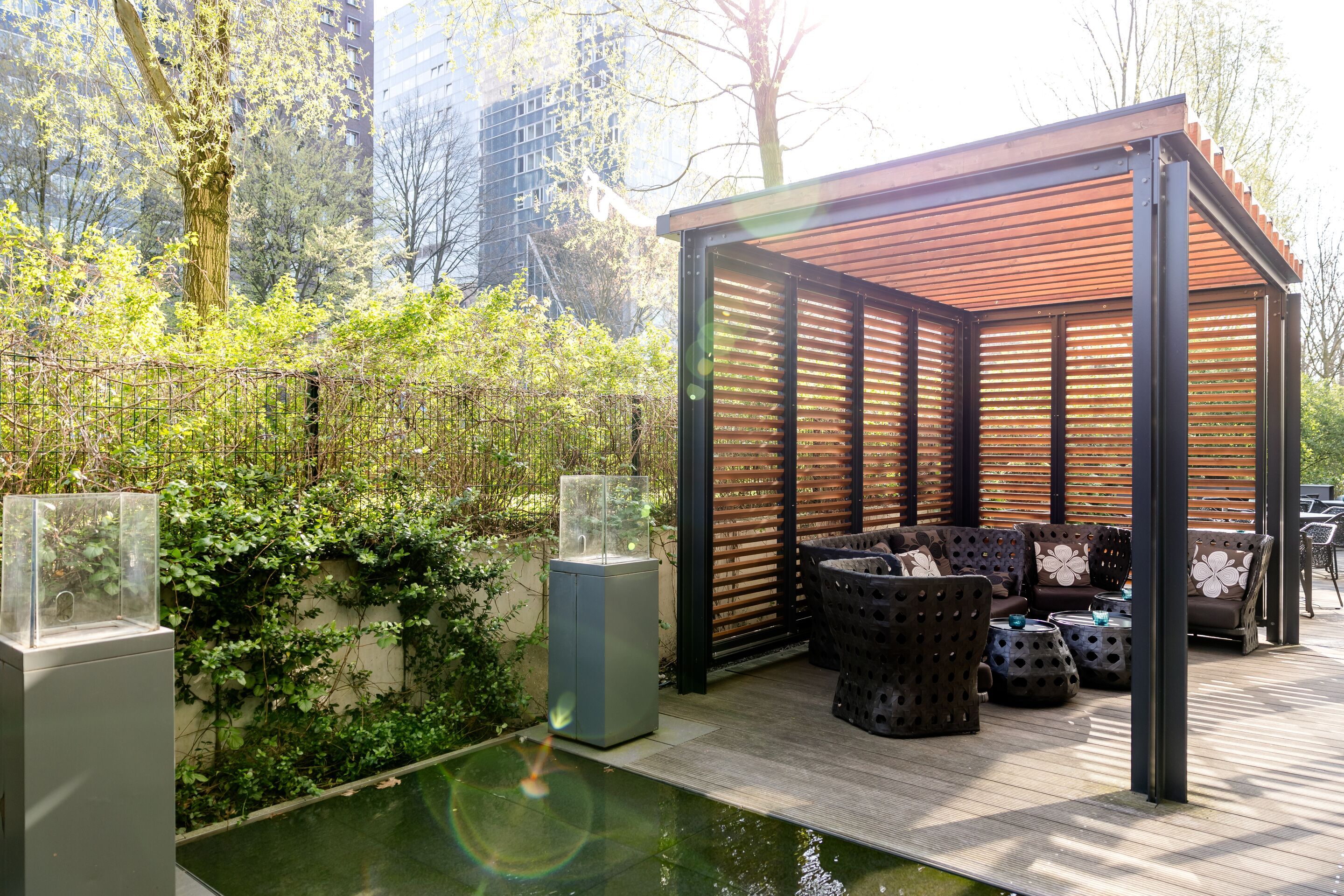 Hotel Hamburg-Alster Motel One geschützter Lounge-Bereich im Freien mit moderner Pergola, Korbsesseln und kleinen Tischen, umgeben von grünem Pflanzenbewuchs und Sonnenschein.