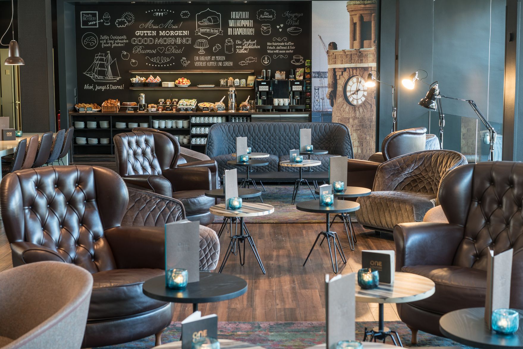 Hotel Hamburg Altona Motel One helle Lounge mit Sesseln sowie Frühstücksbuffet im Hintergrund