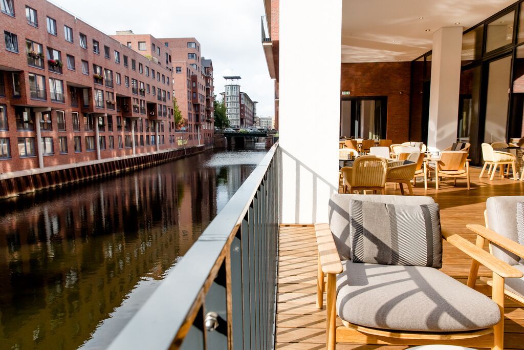 Balkon mit Holzstühlen und gemütlicher Lounge, Blick auf ruhigen Kanal und moderne Backsteingebäude, direkt neben einem eleganten Restaurant mit großen Fenstern. Atmosphäre lädt zum Entspannen und Genießen der Wasserlage ein.