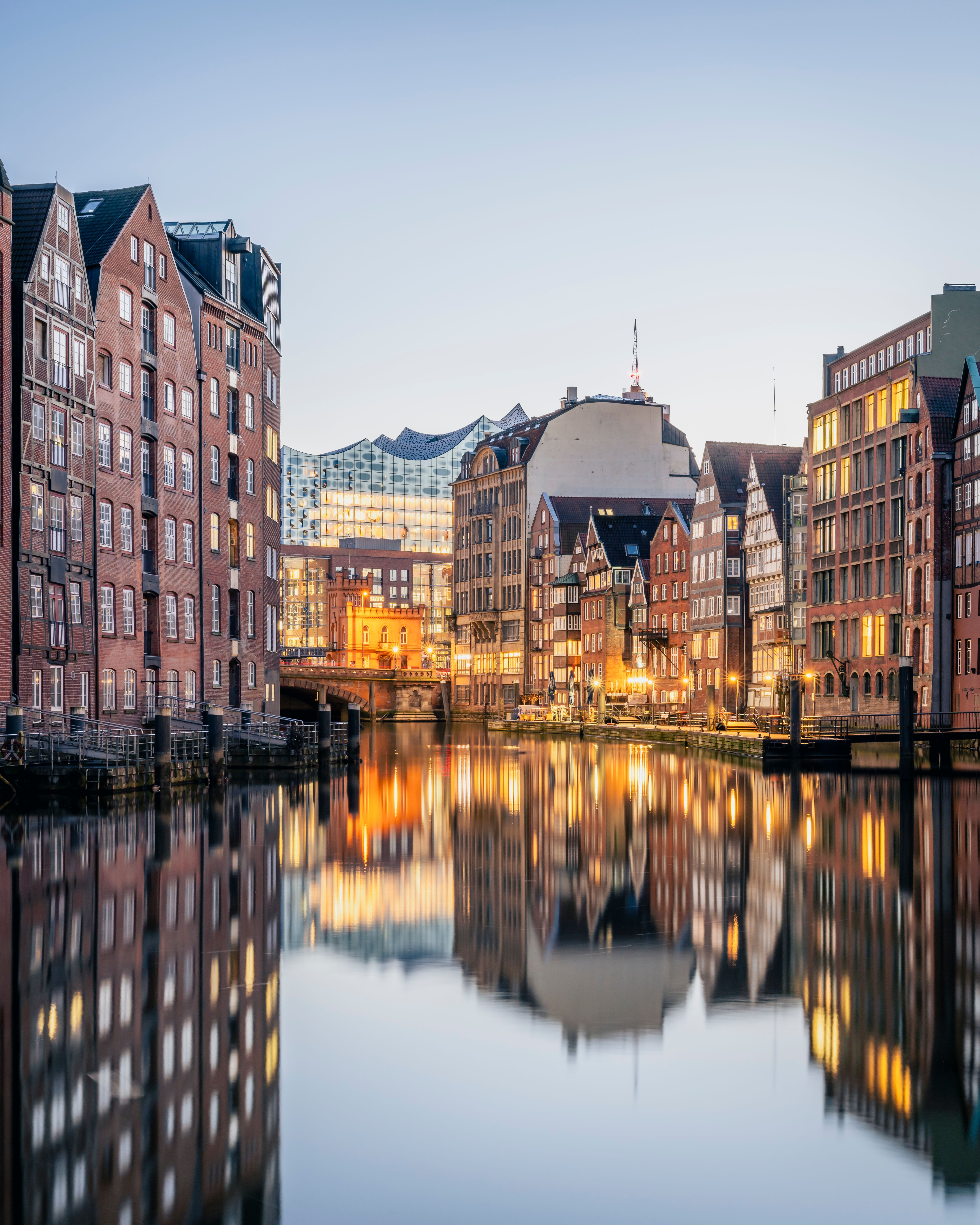 Abendliche Stadtansicht mit historischen Backsteinhäusern und moderner Architektur entlang eines ruhigen Kanals, dessen Wasser die beleuchteten Gebäude spiegeln. Sanfte Beleuchtung schafft eine einladende, atmosphärische Stimmung.