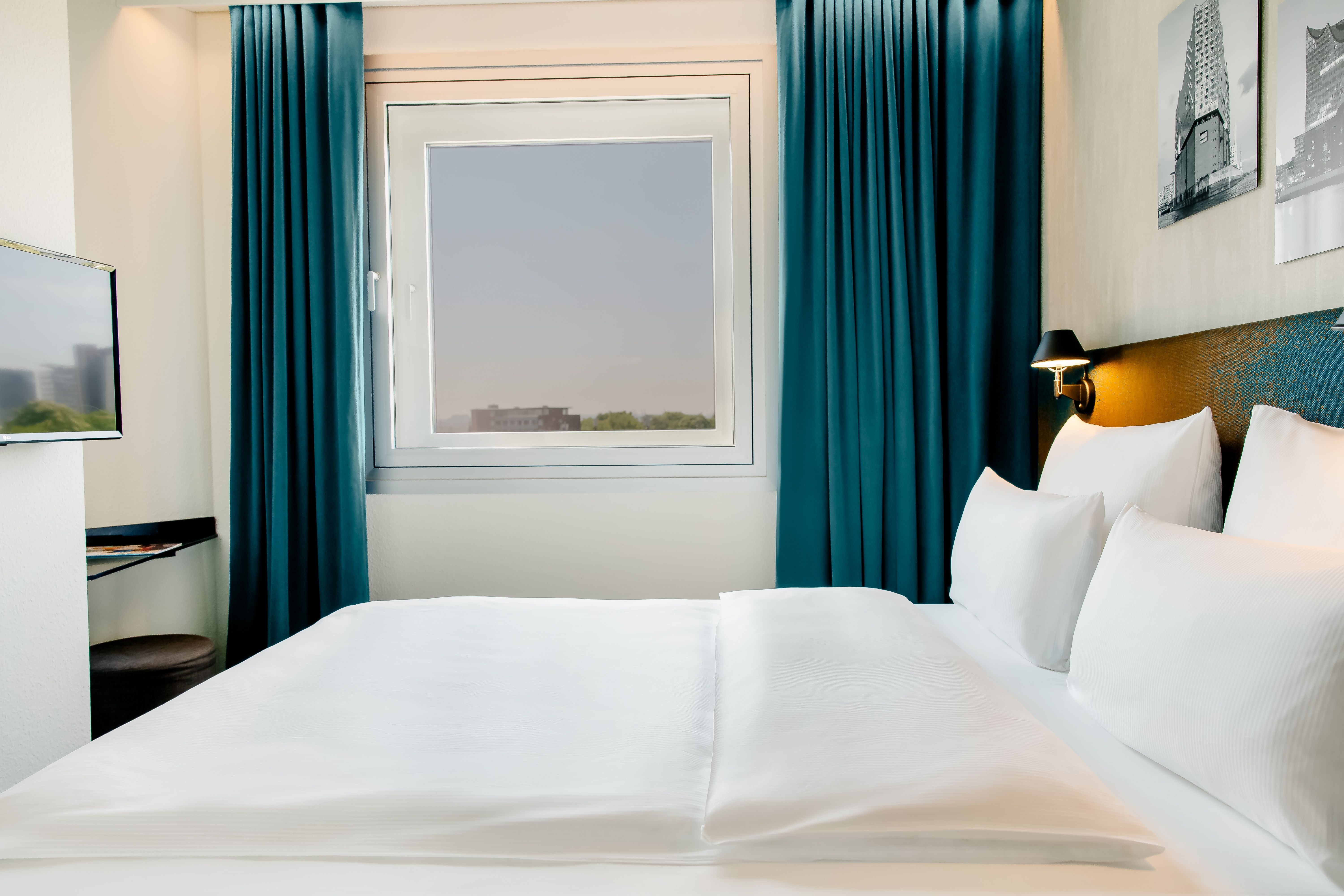 Helles, modernes Hotelzimmer mit großem, bequemem Doppelbett, weißen Bettwäsche, Fenster mit Blick nach draußen und dunkelblauen Vorhängen, das eine gemütliche und ruhige Atmosphäre für erholsamen Aufenthalt schafft.