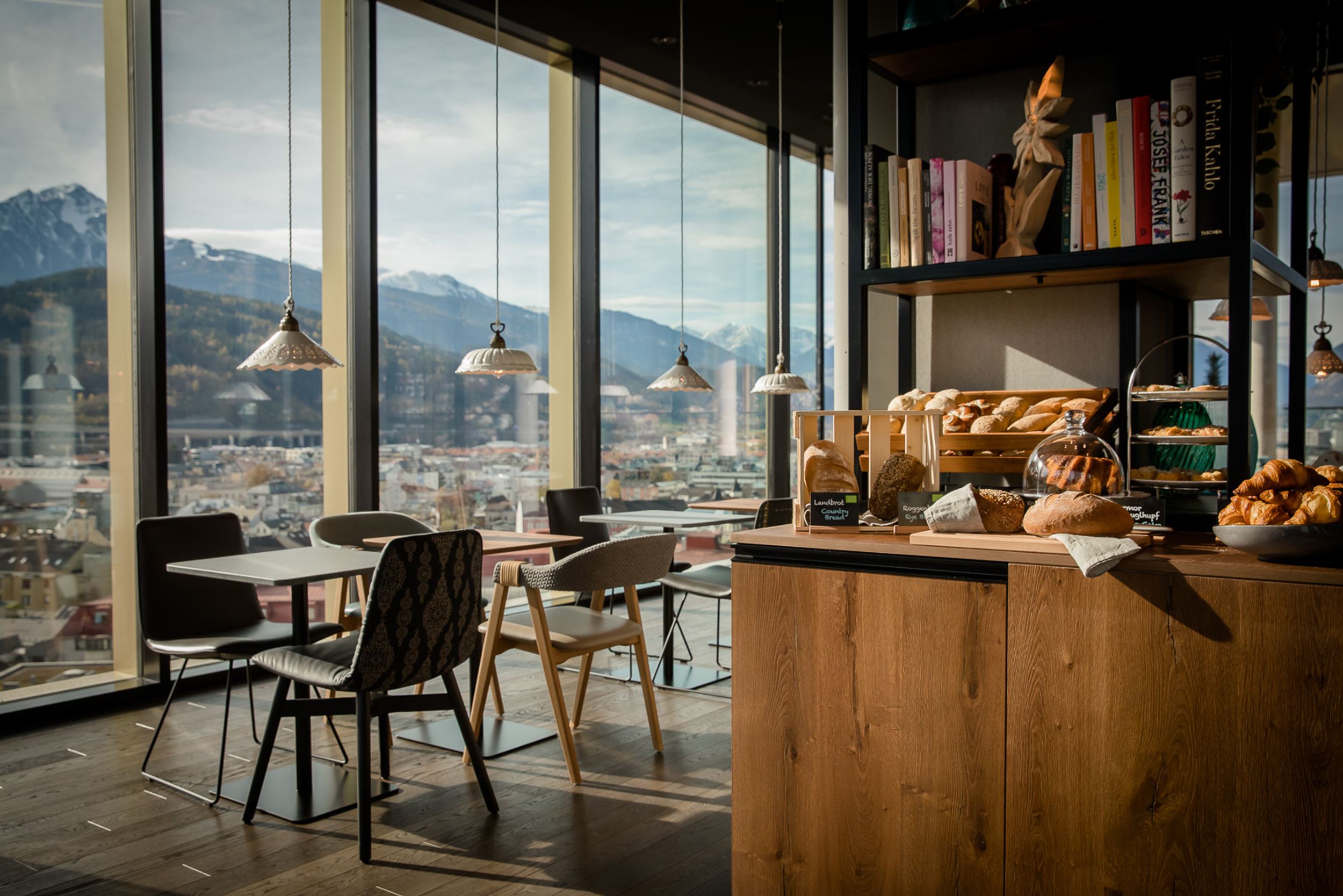 Designhotel Innsbruck Motel One Frühstücksbereich mit mehreren Sitzgelegenheiten und bodentiefen Fenstern mit Sicht auf Berge und Stadt
