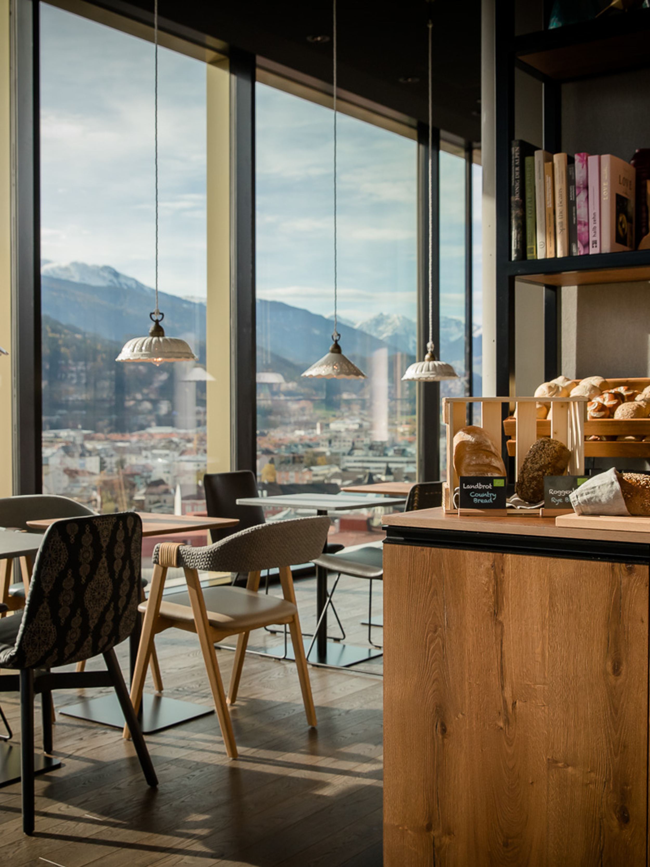Designhotel Innsbruck Motel One Frühstücksbereich mit mehreren Sitzgelegenheiten und bodentiefen Fenstern mit Sicht auf Berge und Stadt