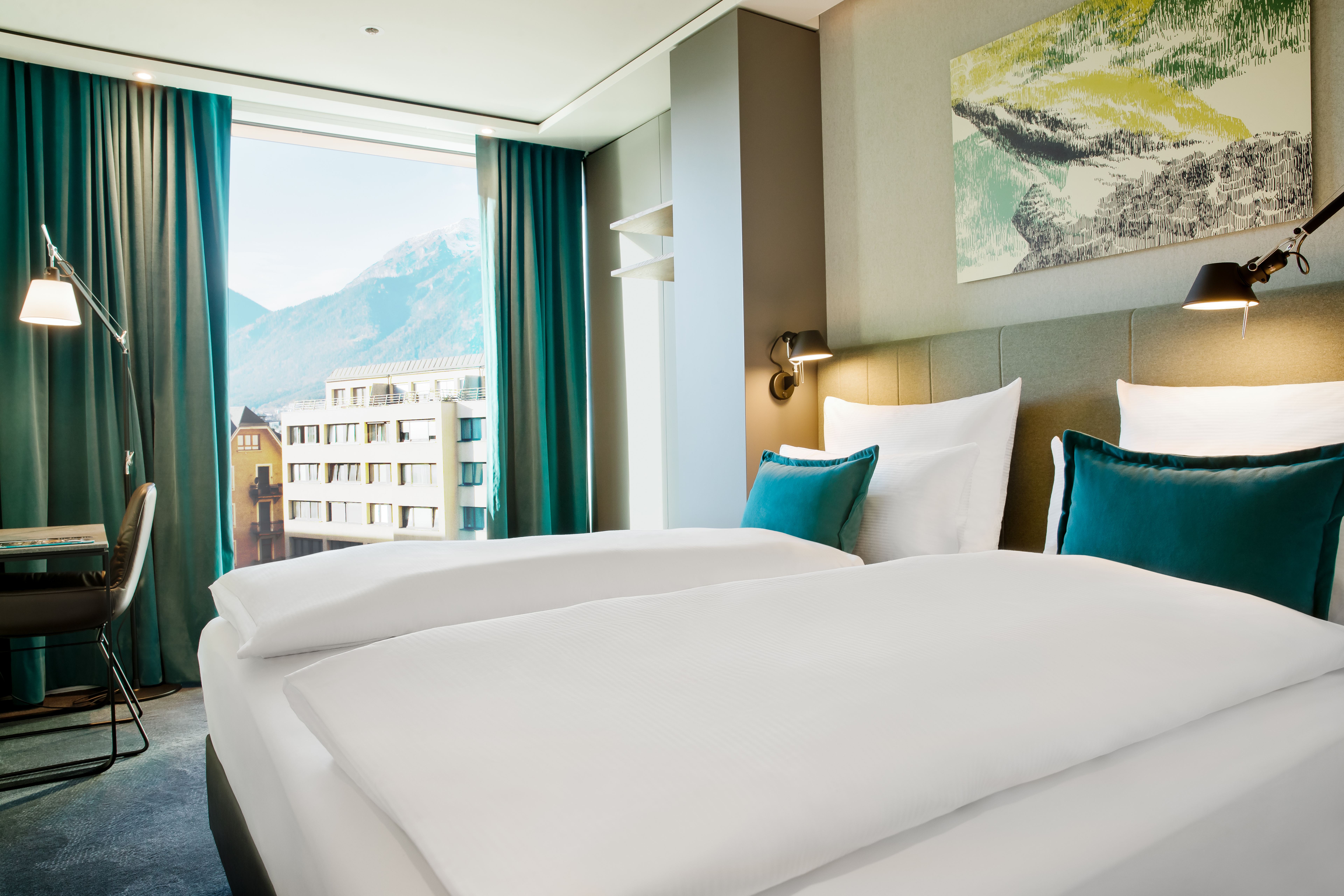Modernes Hotelzimmer mit zwei großen Betten, eleganter Beleuchtung und grün-blauen Akzenten, davor großes Fenster mit Blick auf Berglandschaft und städtische Gebäude. Modernes Hotelzimmer mit zwei großen Betten, eleganter Beleuchtung und grün-blauen Akzenten, davor großes Fenster mit Blick auf Berglandschaft und städtische Gebäude.