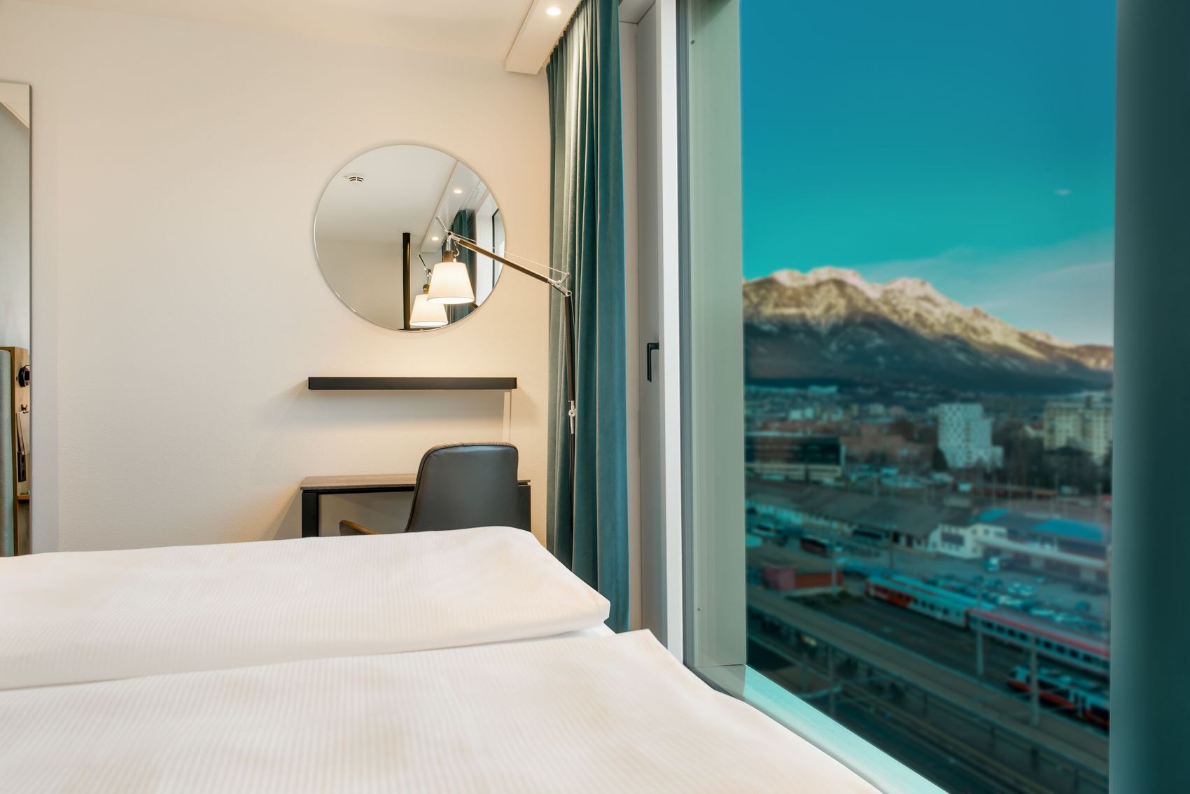 Hotel Motel One Innsbruck Hell eingerichtetes Hotelzimmer mit zwei Betten, Schreibtisch und Stuhl. Große Fenster bieten Ausblick auf eine Stadt und Berge bei klarem Himmel.