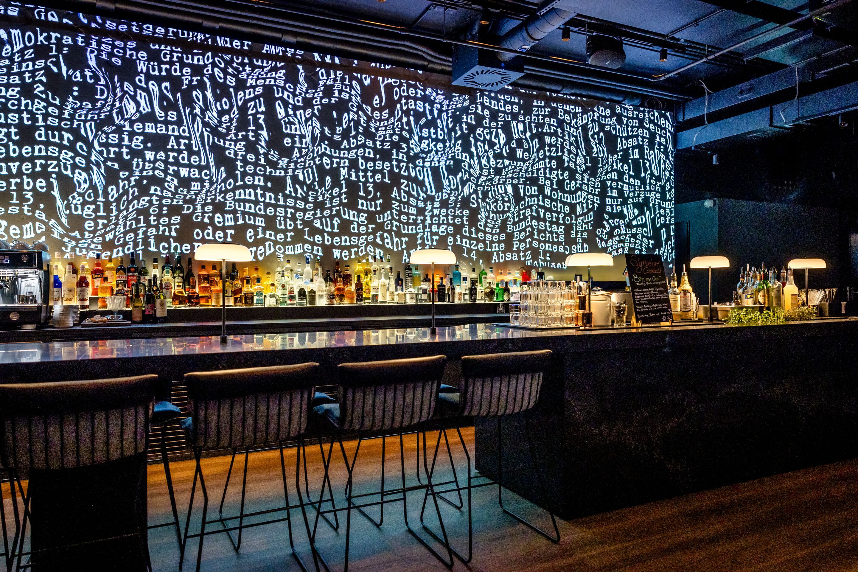 Hotel Karlsruhe Motel One Rooftop Bar mit beleuchtetem Tresen und hohen Barstühlen. Die Wand hinter der Bar zeigt eine großflächige Projektion aus weißem Text auf schwarzem Hintergrund.