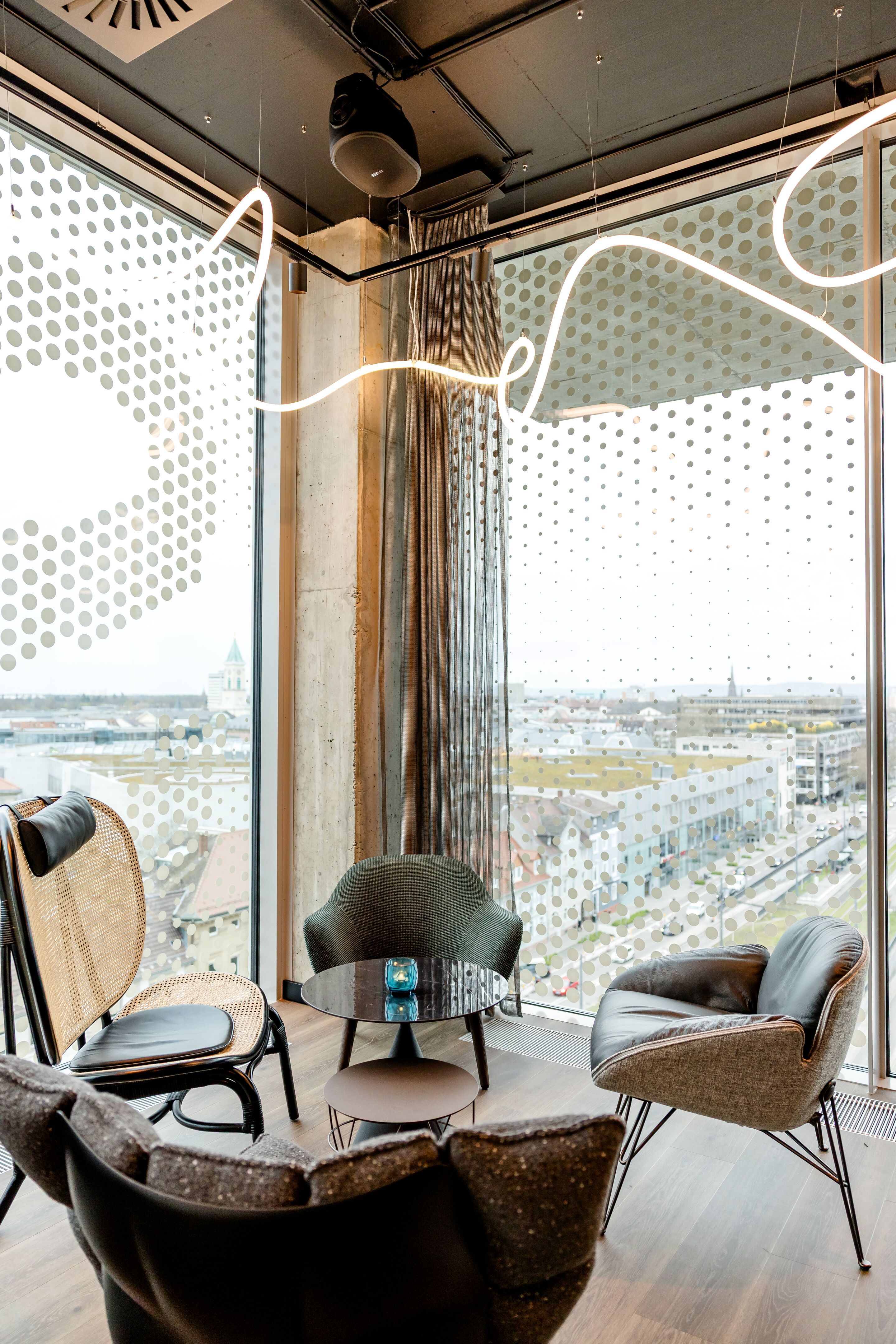Hotel Karlsruhe Motel One Rooftop Bar Sitzecke mit modernen Sesseln und kleinem Tisch, umgeben von großen Panoramafenstern mit Blick auf die Stadt.
