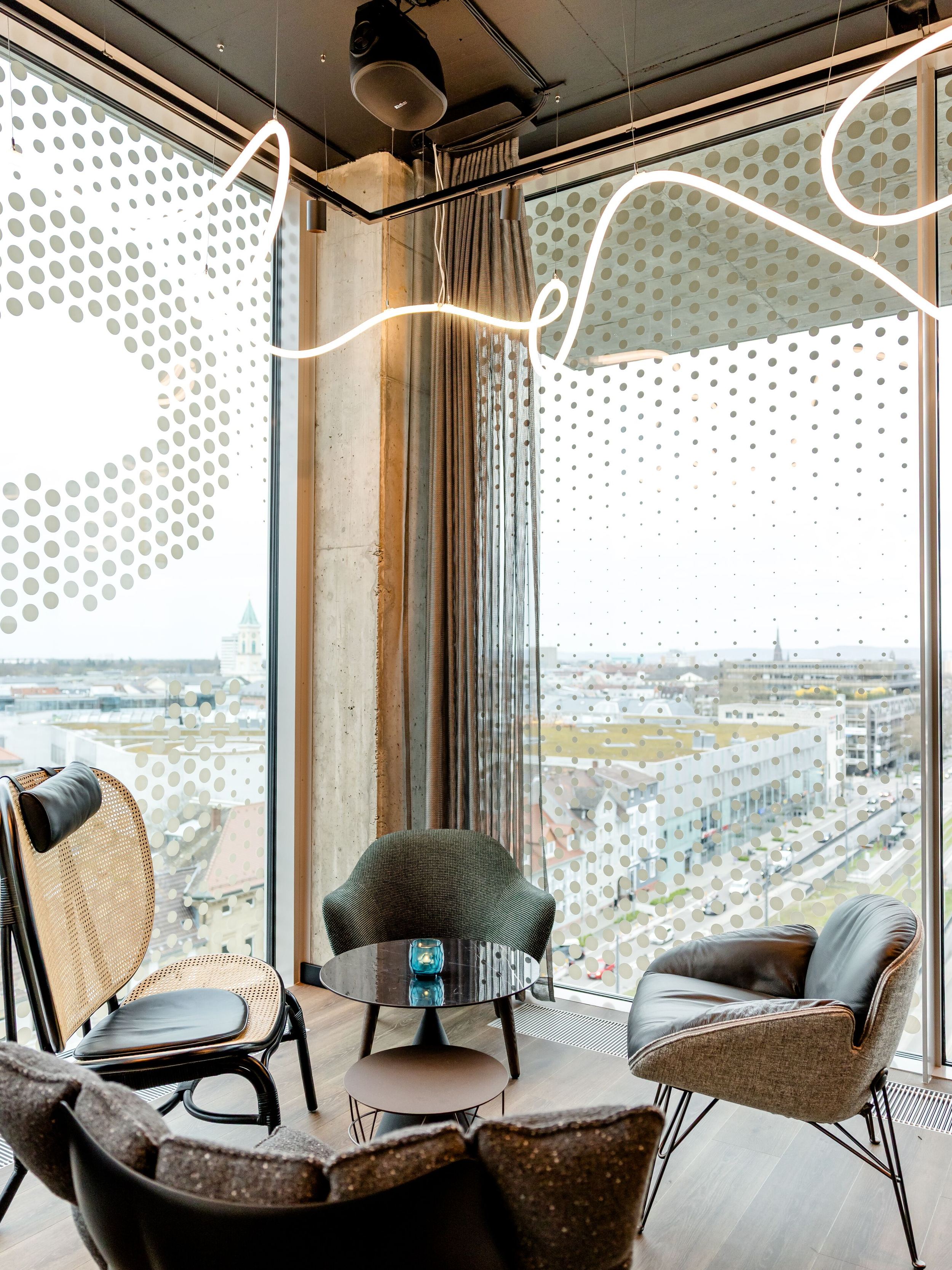 Hotel Karlsruhe Motel One Rooftop Bar Sitzecke mit modernen Sesseln und kleinem Tisch, umgeben von großen Panoramafenstern mit Blick auf die Stadt.